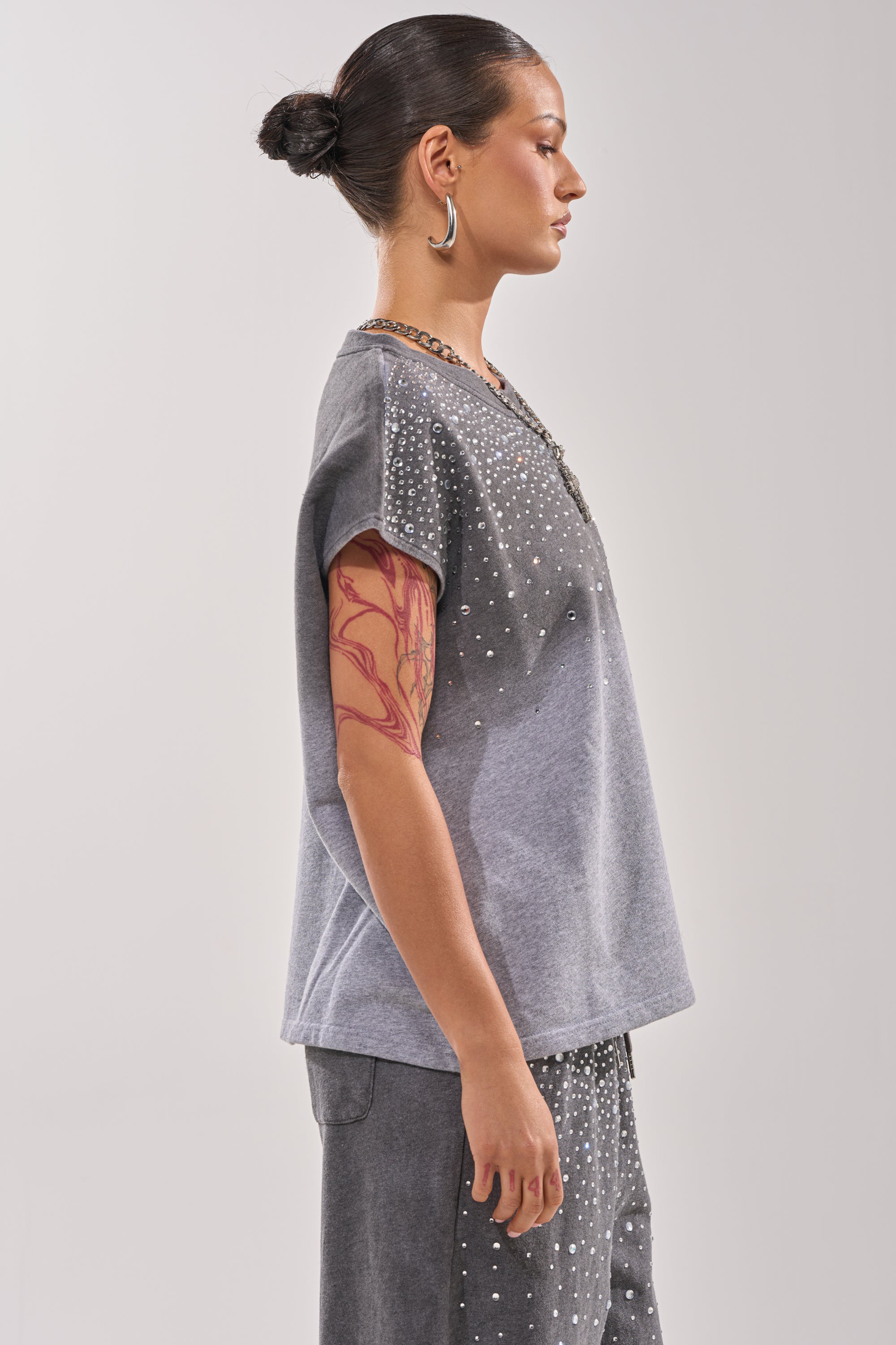 A woman with her hair in a bun, wearing the NIGHT SKY RHINESTONE TEE and matching studded pants, stands in profile. She has large hoop earrings and a red tattoo on her upper arm. 2