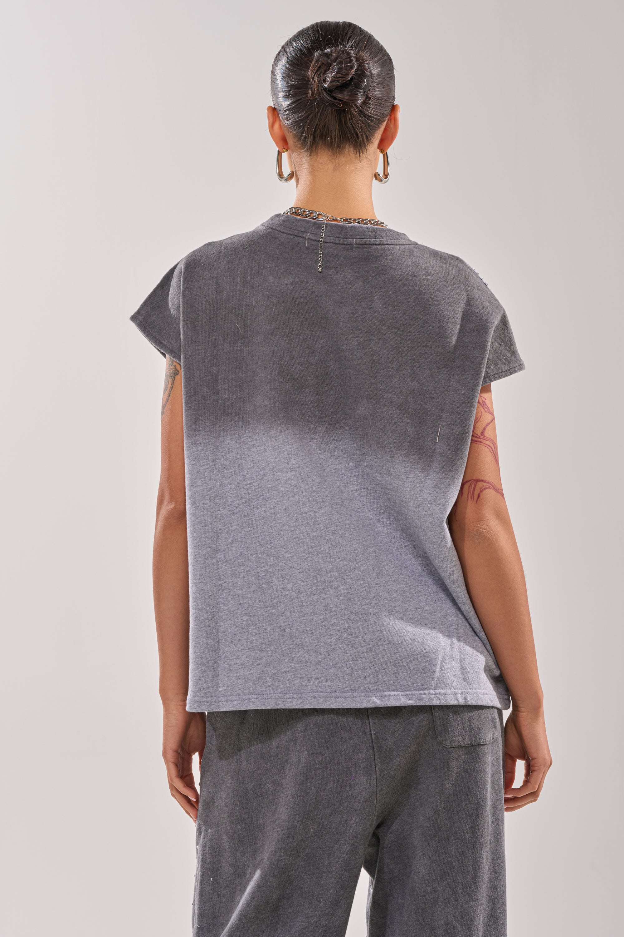 A person with their hair in a bun, wearing the NIGHT SKY RHINESTONE TEE and matching gray pants, stands with their back to the camera. Hoop earrings and tattoos on their left arm are visible. 3