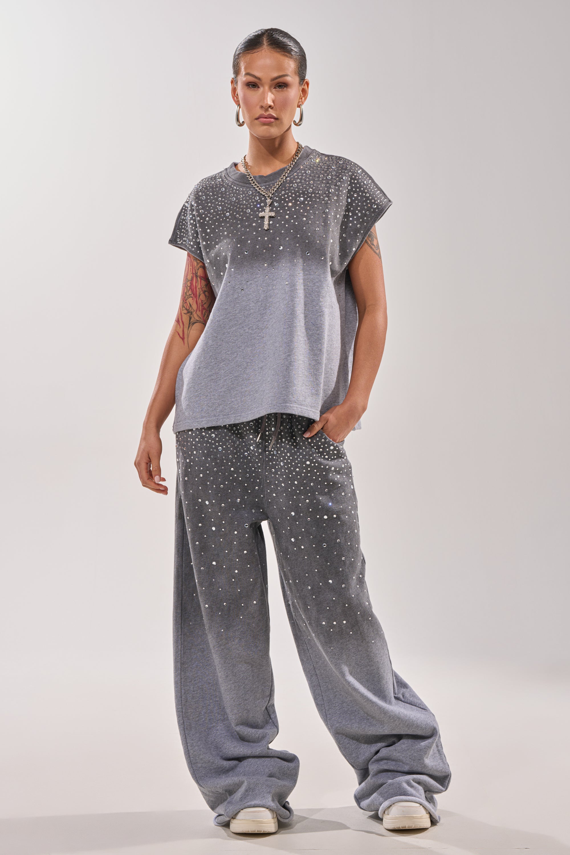 A person stands confidently in the NIGHT SKY RHINESTONE TEE and matching wide-leg pants with white sneakers, slicked-back hair, visible tattoos, hoop earrings, and a cross necklace. 4