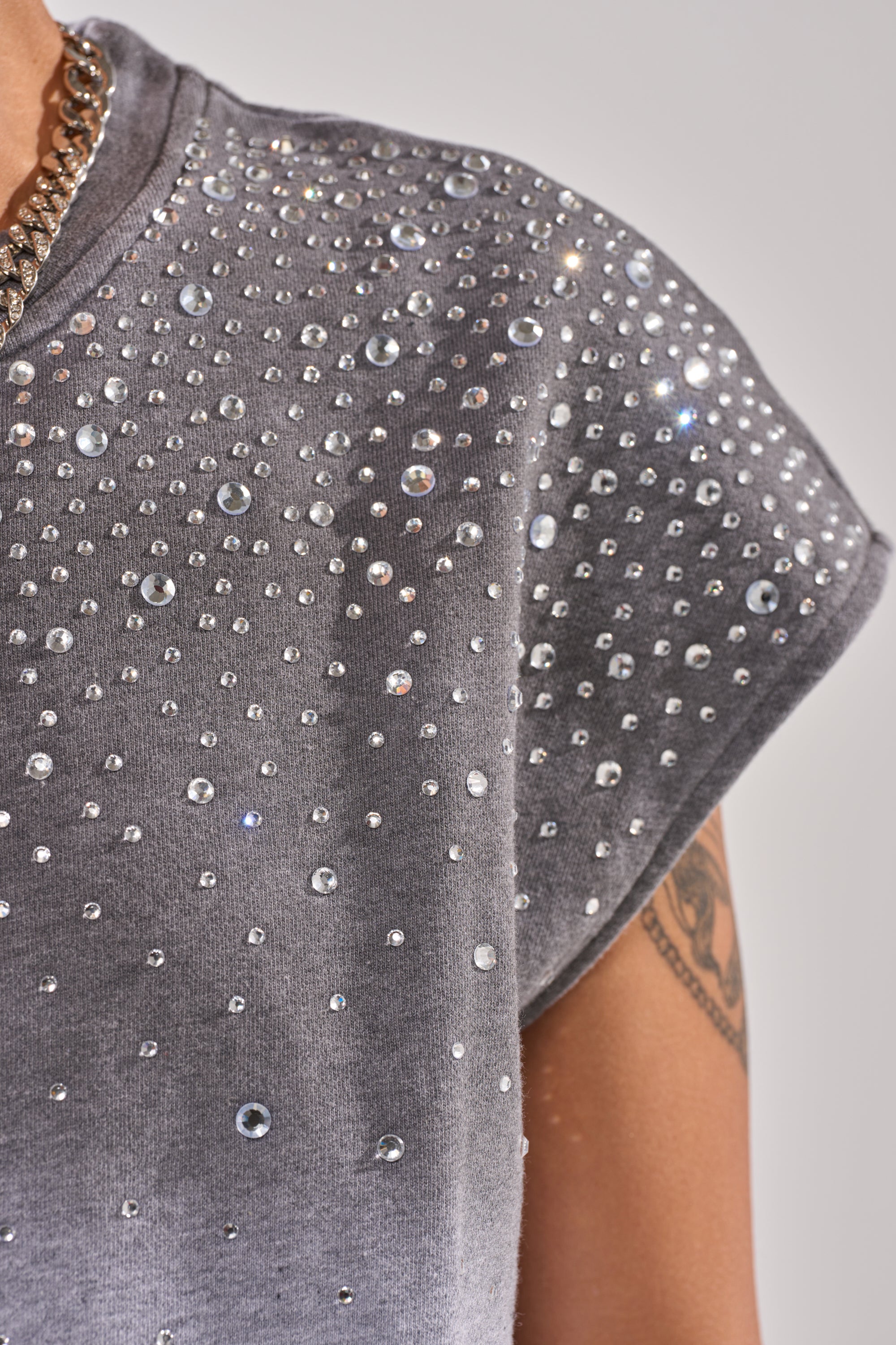 A close-up of a person's shoulder in the NIGHT SKY RHINESTONE TEE, grey casual wear sparkling with rhinestones. A chain necklace and upper arm tattoo are visible against a plain background. 6