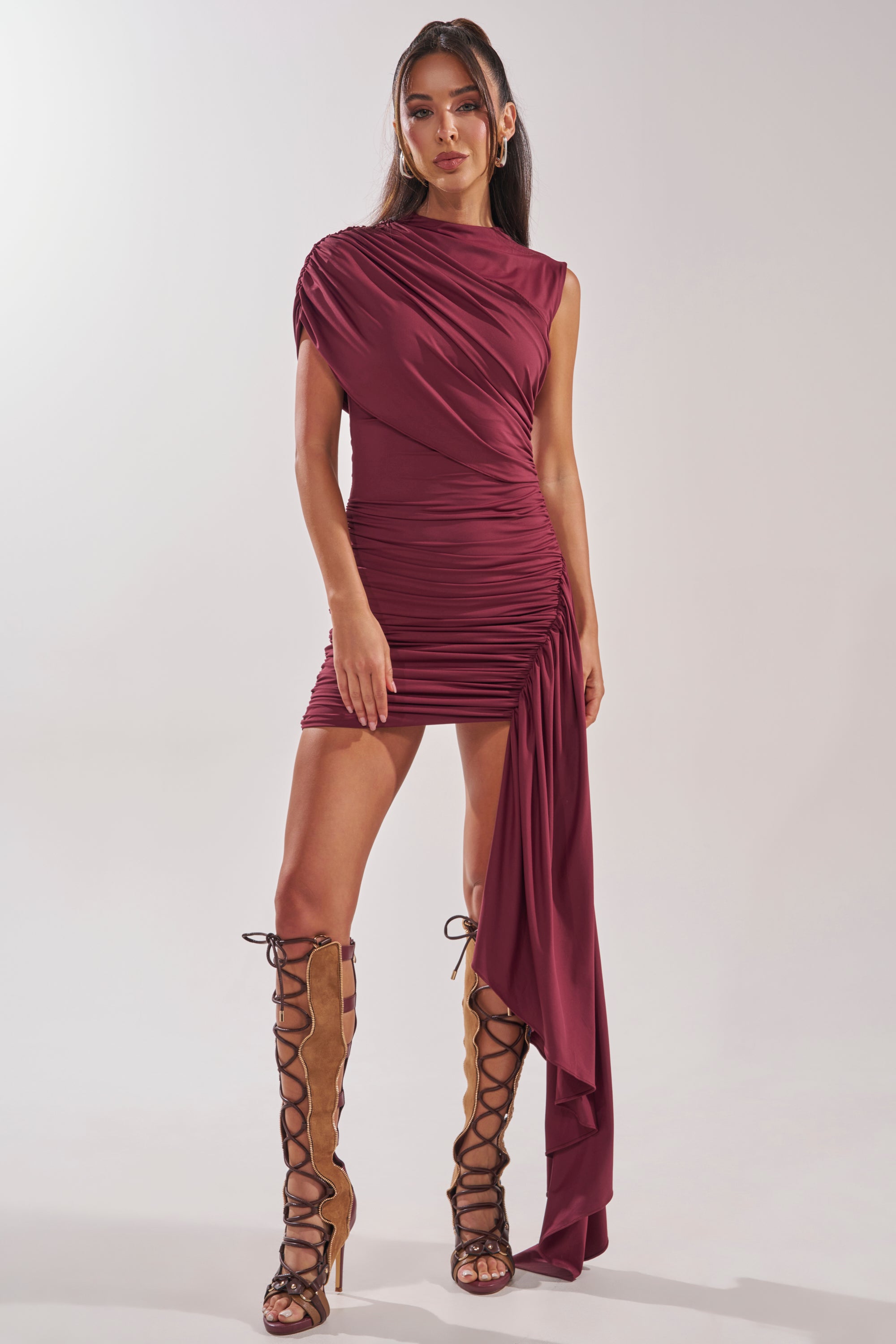 Woman wearing the WRAP ME UP MINI DRESS—burgundy, asymmetrical, with one-shoulder ruching and drape—paired with tan lace-up knee-high boots; plain light gray background. 1
