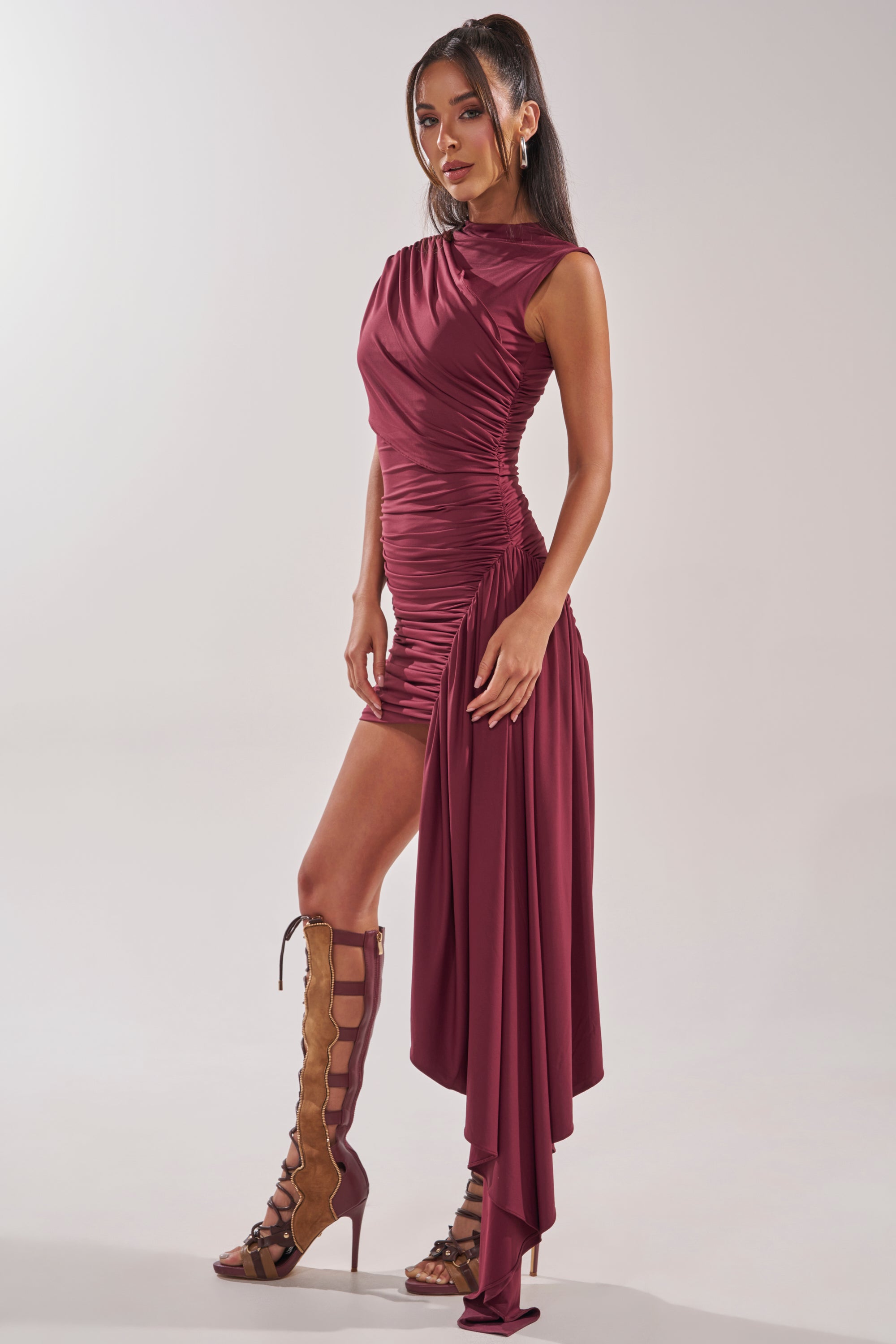 A woman poses against a plain light background wearing the WRAP ME UP MINI DRESS, a sleeveless burgundy mini with a ruched bodice and asymmetric draped skirt, styled with tan lace-up knee-high boots. 4