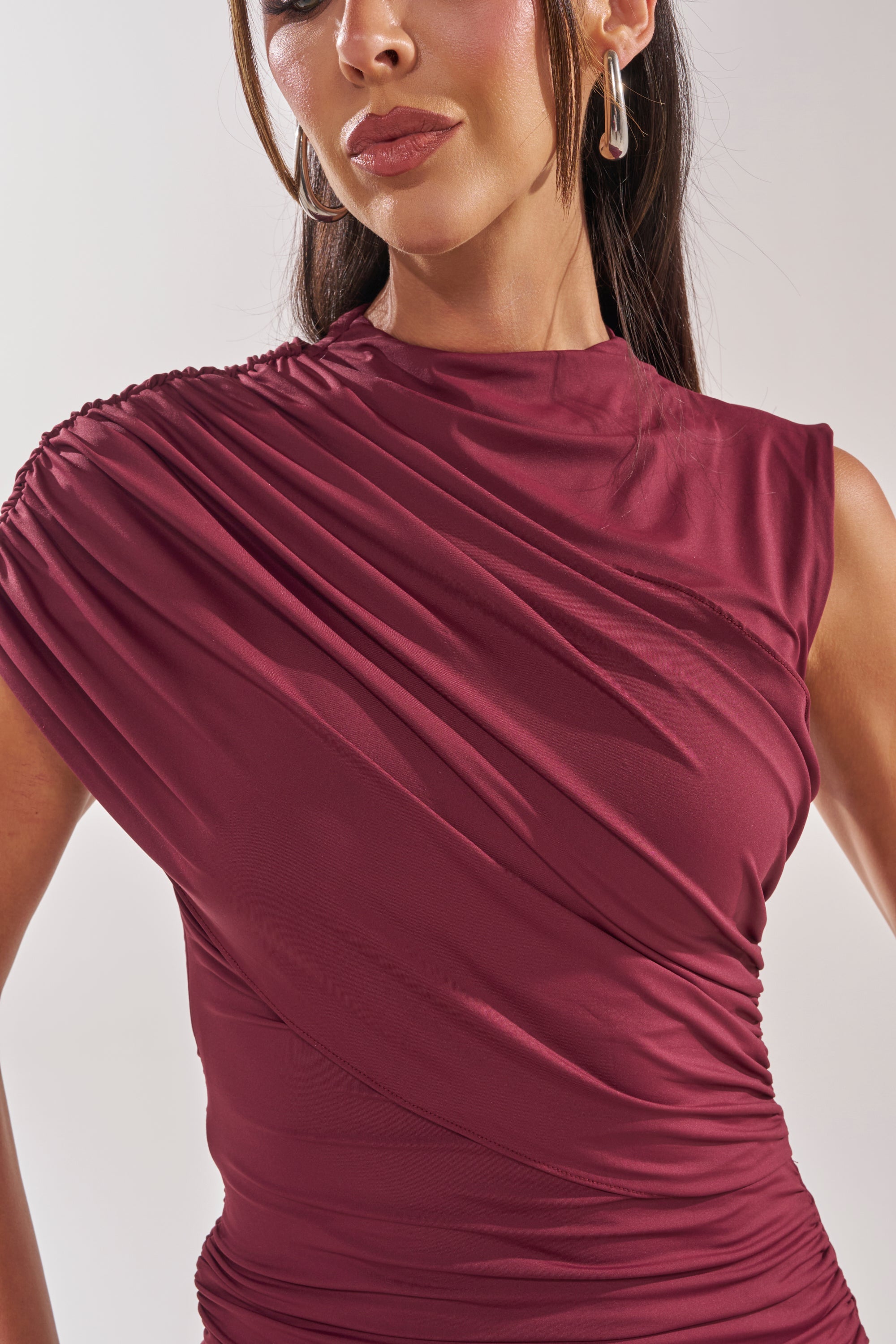 A woman wears the WRAP ME UP MINI DRESS, a sleeveless maroon mini with ruched and draped one-shoulder detailing. She accessorizes with large hoop earrings; her face is partly visible against a light, plain background. 7