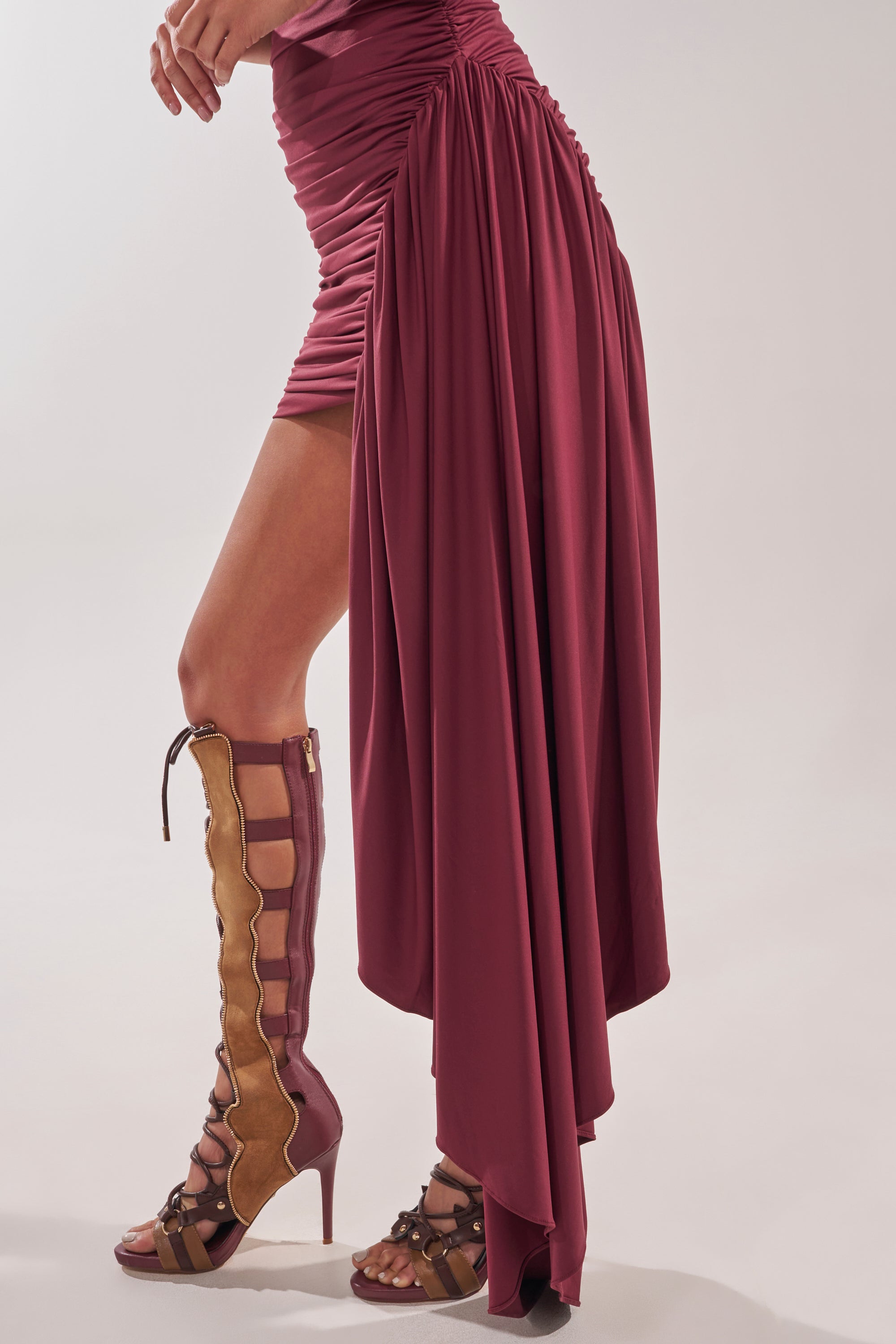 Someone wearing the WRAP ME UP MINI DRESS—an asymmetrical, ruched burgundy mini dress with a dramatic draped train—and brown lace-up cut-out high-heeled boots, standing against a neutral background. 8
