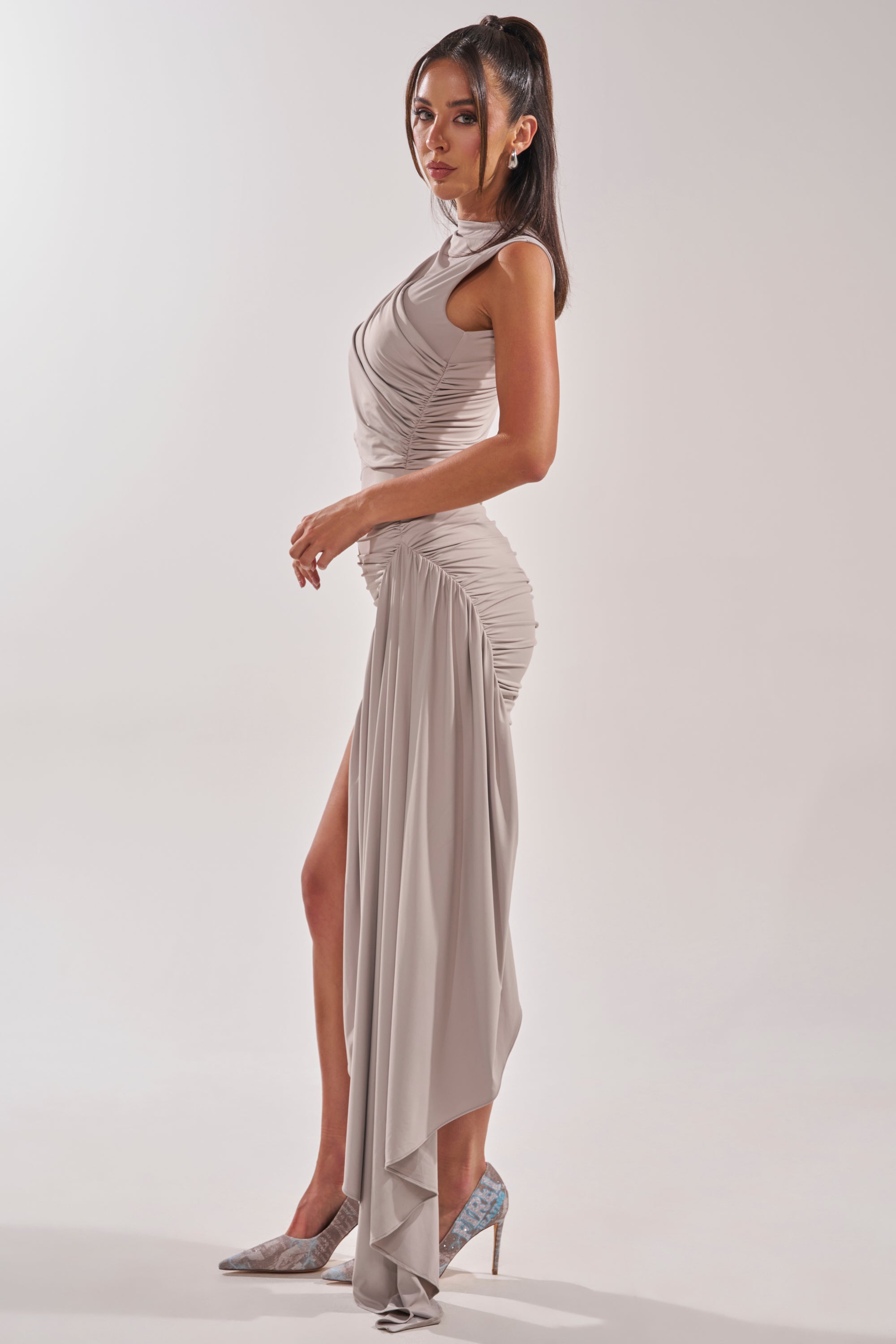 A woman poses in profile wearing the WRAP ME UP MINI DRESS IN GREY, a ruched cocktail dress with a high slit and draped hem. Her long dark hair is styled in a high ponytail, paired with sparkly pointed-toe heels against a plain backdrop. 3