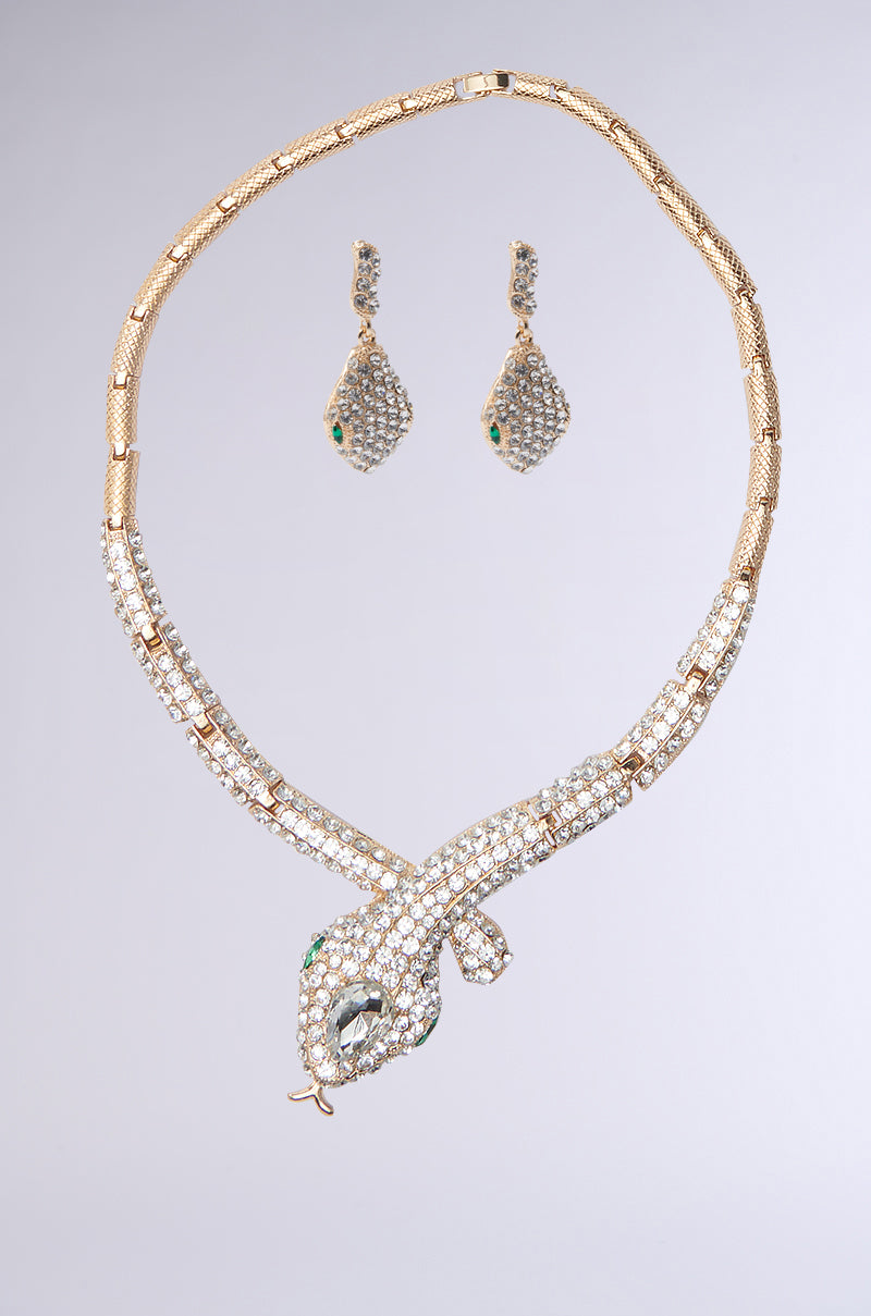 The BITE ME LUXE SNAKE NECKLACE AND EARRINGS SET showcases a gold snake necklace adorned with rhinestones, a diamond-encrusted snake head, and green gemstone eyes. It's complemented by matching teardrop-shaped diamond earrings, all set against a plain gray backdrop.