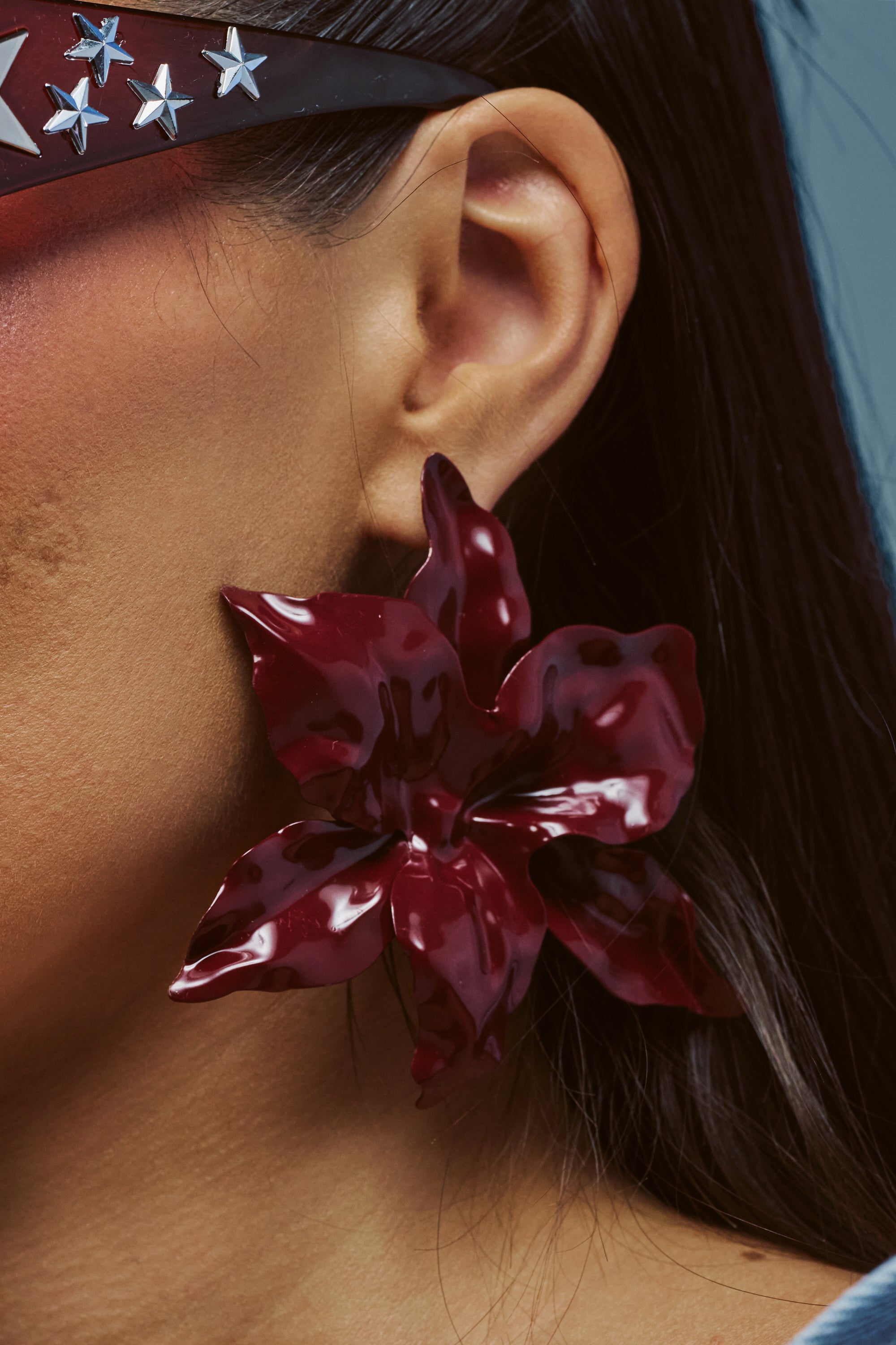 A close-up of a person’s ear wearing the LOVER EARRING—a large, glossy burgundy acrylic lotus flower. The person also wears star-studded sunglasses and has long dark hair.