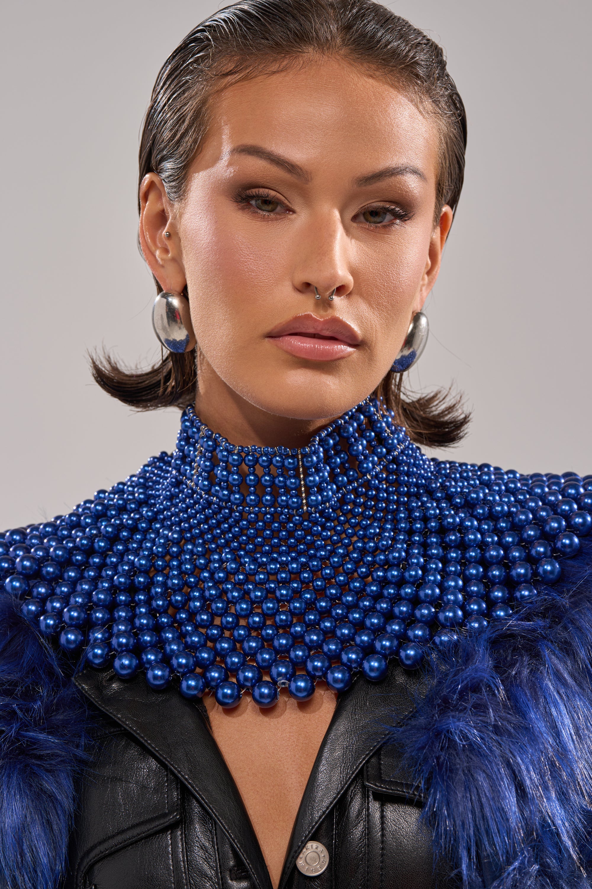 A woman with slicked-back hair wears silver hoop earrings, a black leather top with blue faux fur accents, and the COBALT NECKLACE—a statement collar of layered blue beads—posing against a neutral background.