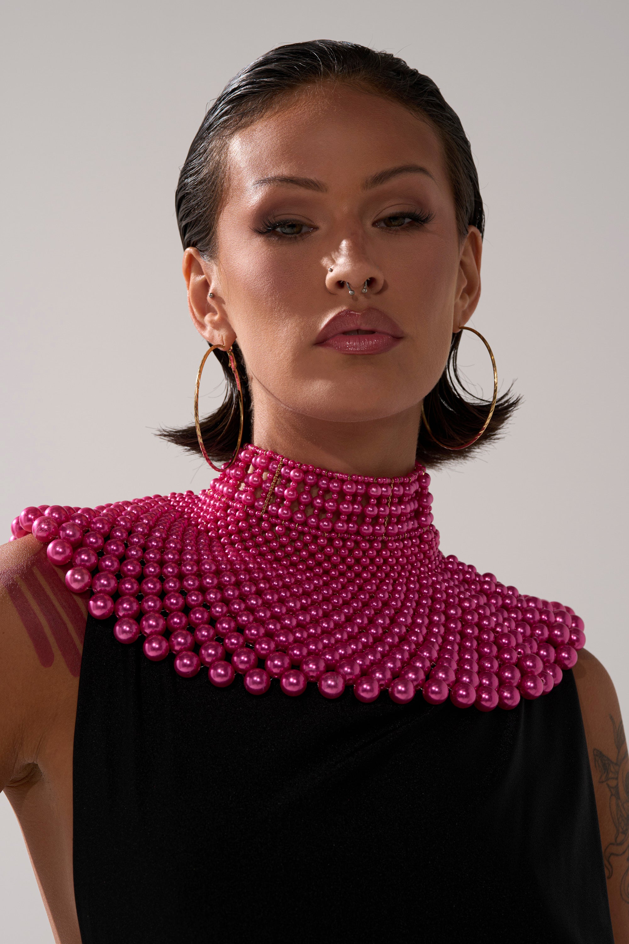A person with slicked-back dark hair, hoop earrings, facial piercings, and a visible arm tattoo wears the high-necked hot pink FEEL MY POWER NECKLACE over a black outfit against a neutral background.