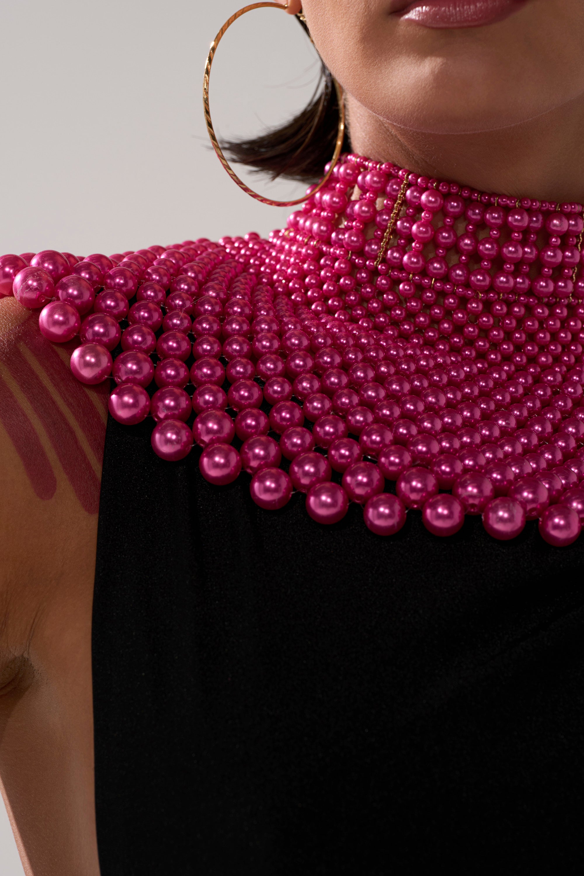 A person wears a sleeveless black top styled with the FEEL MY POWER NECKLACE, a bold layered hot pink beaded statement piece. They accessorize with large gold hoops, glossy pink lips, and have pink paint streaks on their upper arm.