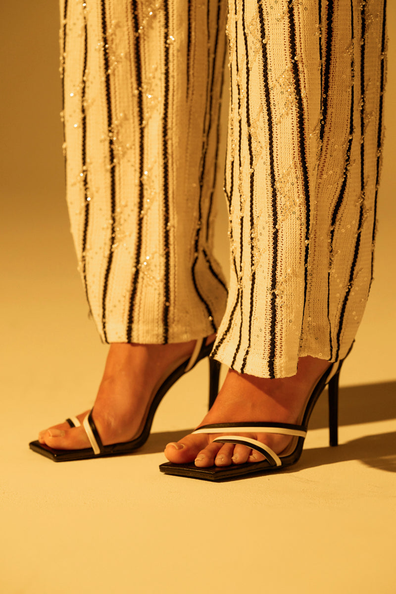 A person wears the AKIRA NEHARA BLACK AND WHITE STILETTO SANDAL, crafted from vegan leather with thin white straps and an open square toe. They pair it with sequined pants featuring vertical stripes. The softly lit background adds a warm tone.