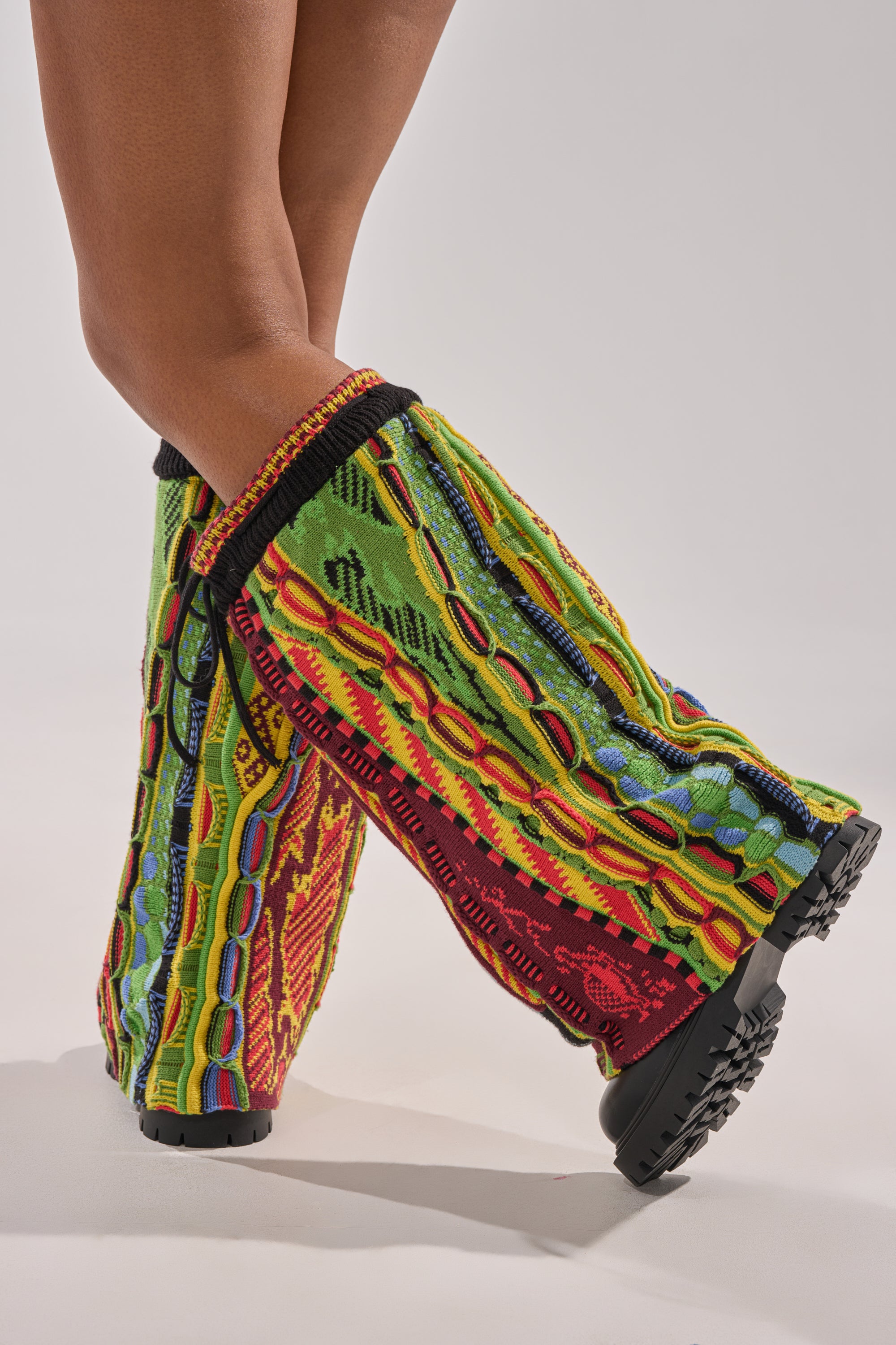 A person stands with legs crossed, wearing tall, colorful patterned leg warmers over the AKIRA GOLD NEIL SWEATER COMBAT BOOTIE. The leg warmers are bright green, red, yellow, and blue. The background is plain and light-colored.
