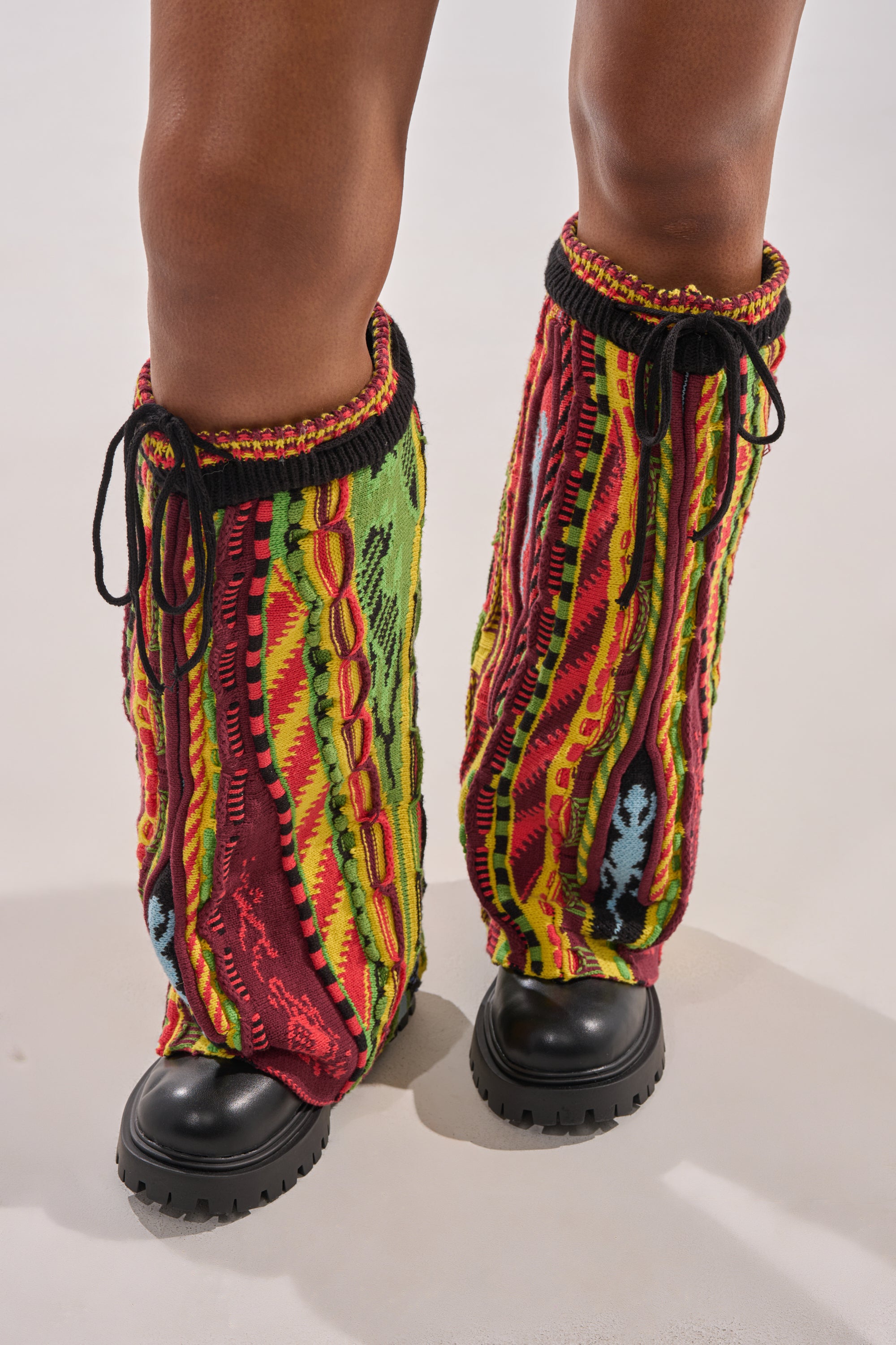 A person wears the AKIRA GOLD NEIL SWEATER COMBAT BOOTIE—chunky heel faux leather boots styled with colorful, knit leg warmers featuring red, yellow, green, and black patterns—standing on a light background.