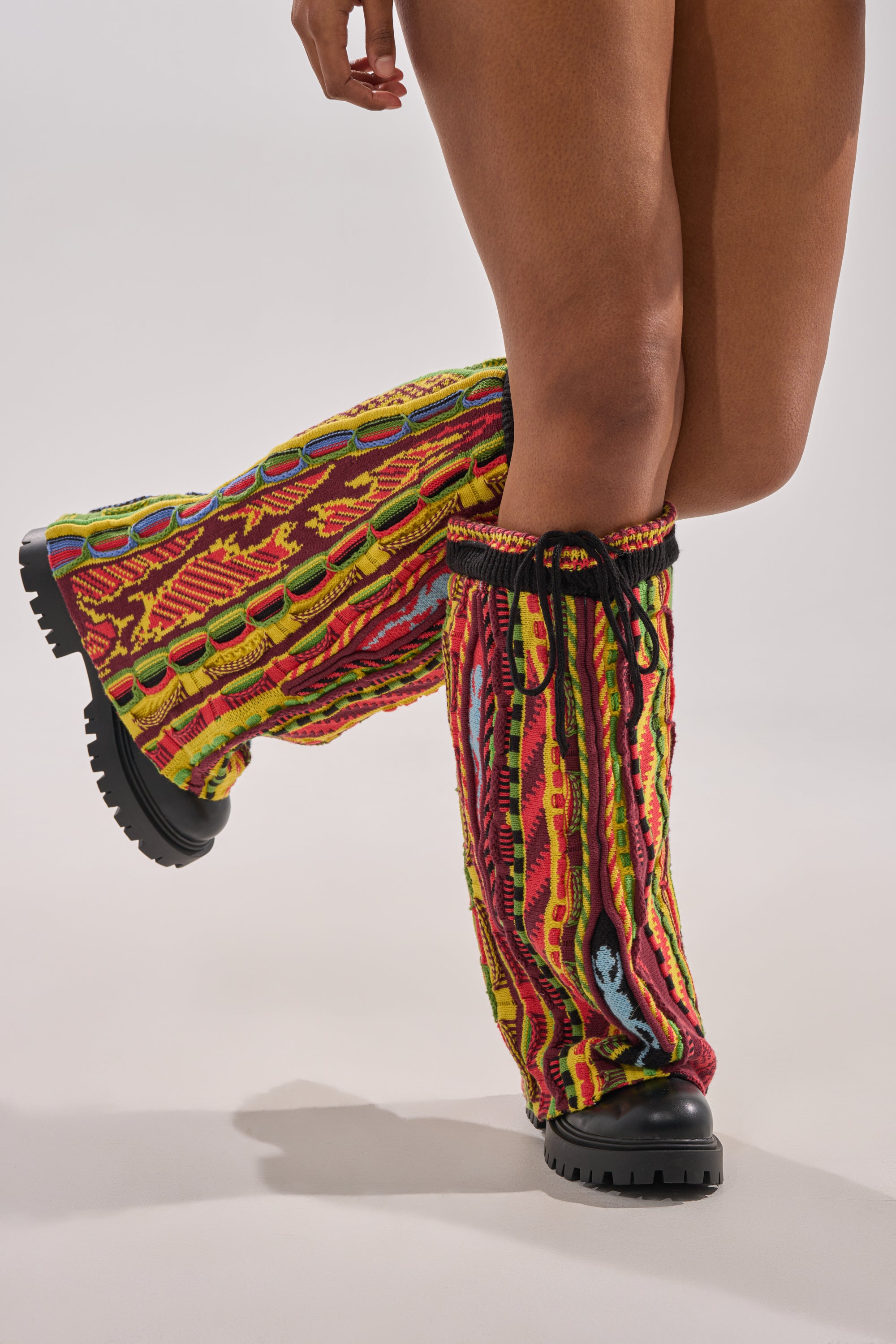 Wearing AKIRA GOLD NEIL SWEATER COMBAT BOOTIE, a person stands on one leg with the other bent at the knee, pairing chunky black heel boots and patterned leg warmers against a plain light background.