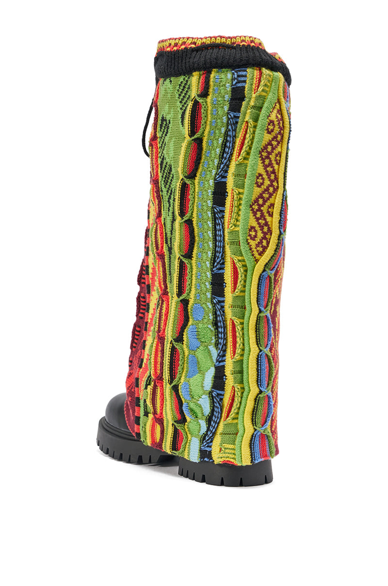 The AKIRA GOLD NEIL SWEATER COMBAT BOOTIE is a tall boot featuring a chunky black lug sole and a colorful textured knit upper in green, red, yellow, blue, and orange patterns reminiscent of a vintage sweater.