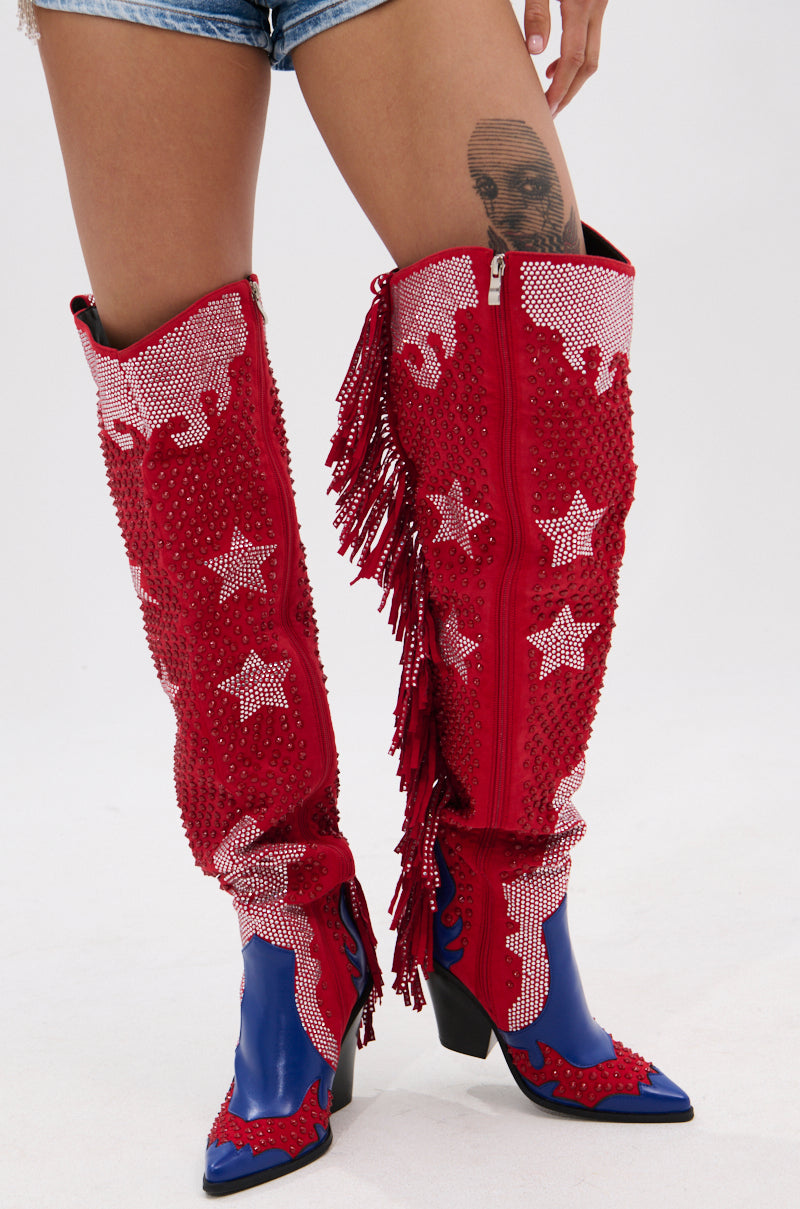 Someone wearing denim shorts and showing a thigh tattoo styles the AZALEA WANG NELLA BLUE MULTI WESTERN EMBELLISHED BOOT—tall red faux suede boots with white stars, fringe, and rhinestone accents on blue pointed toes and heels.
