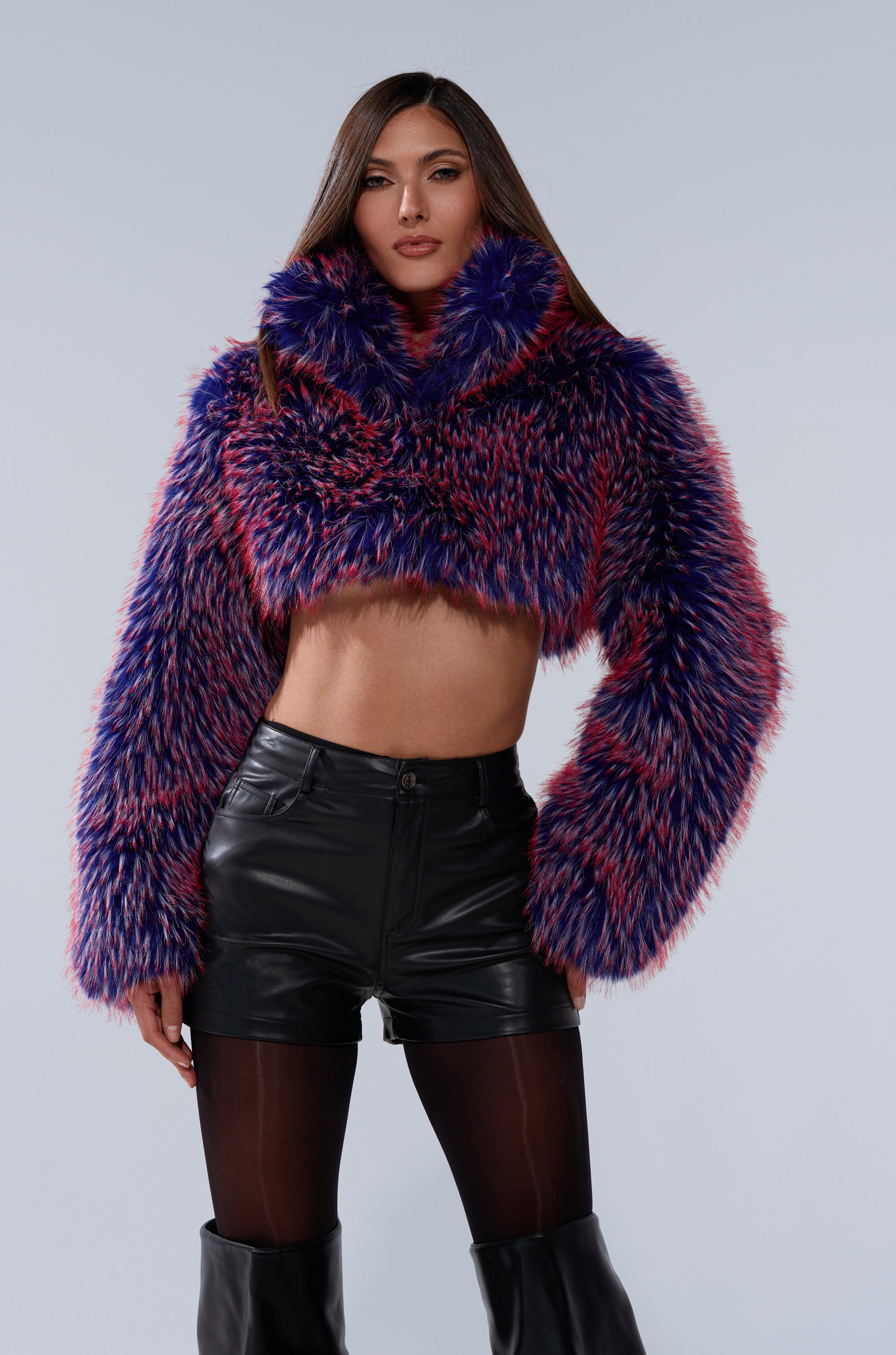 A woman stands confidently in the NELLIE FAUX FUR BOLERO, styled with black high-waisted shorts, sheer black tights, and tall black boots against a plain light background.