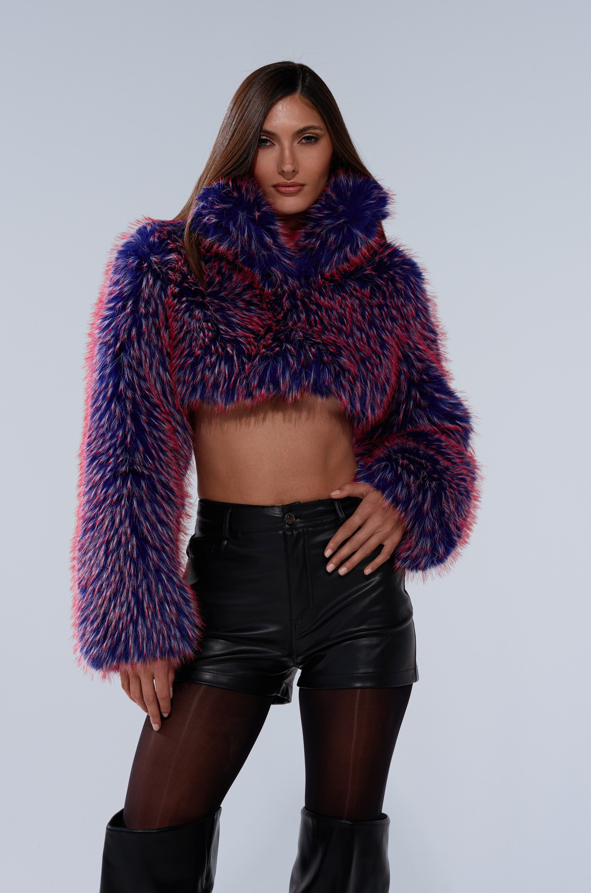 A woman poses confidently against a plain light background wearing the NELLIE FAUX FUR BOLERO in purple and pink, paired with black leather shorts, black tights, and knee-high black boots.