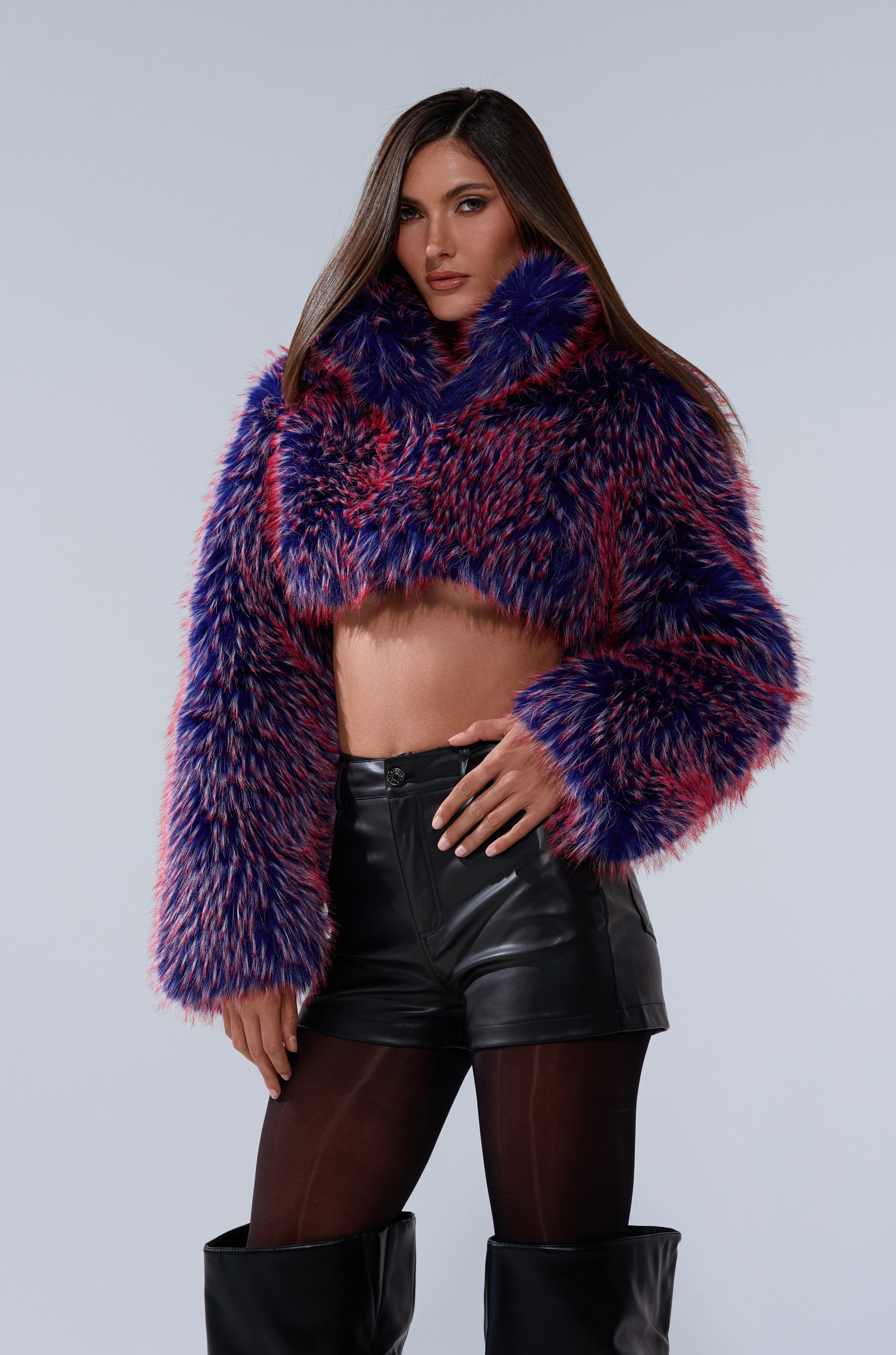 A woman stands confidently against a light-gray background, wearing the NELLIE FAUX FUR BOLERO in vibrant purple and pink, paired with black leather shorts, black tights, and knee-high black boots.