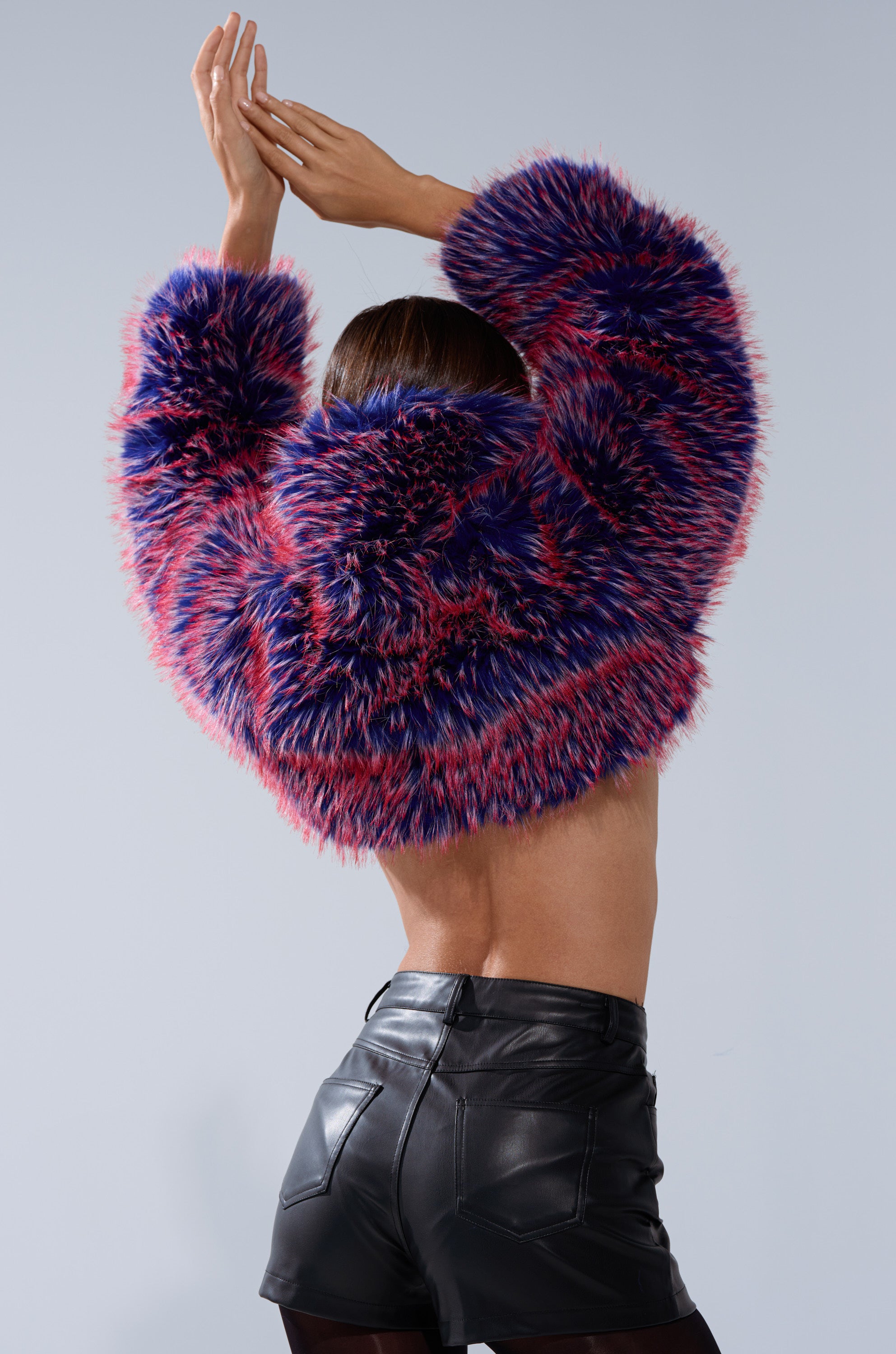 A person faces away, arms raised, wearing the NELLIE FAUX FUR BOLERO in pink and blue shades with high-waisted black leather shorts against a plain background.