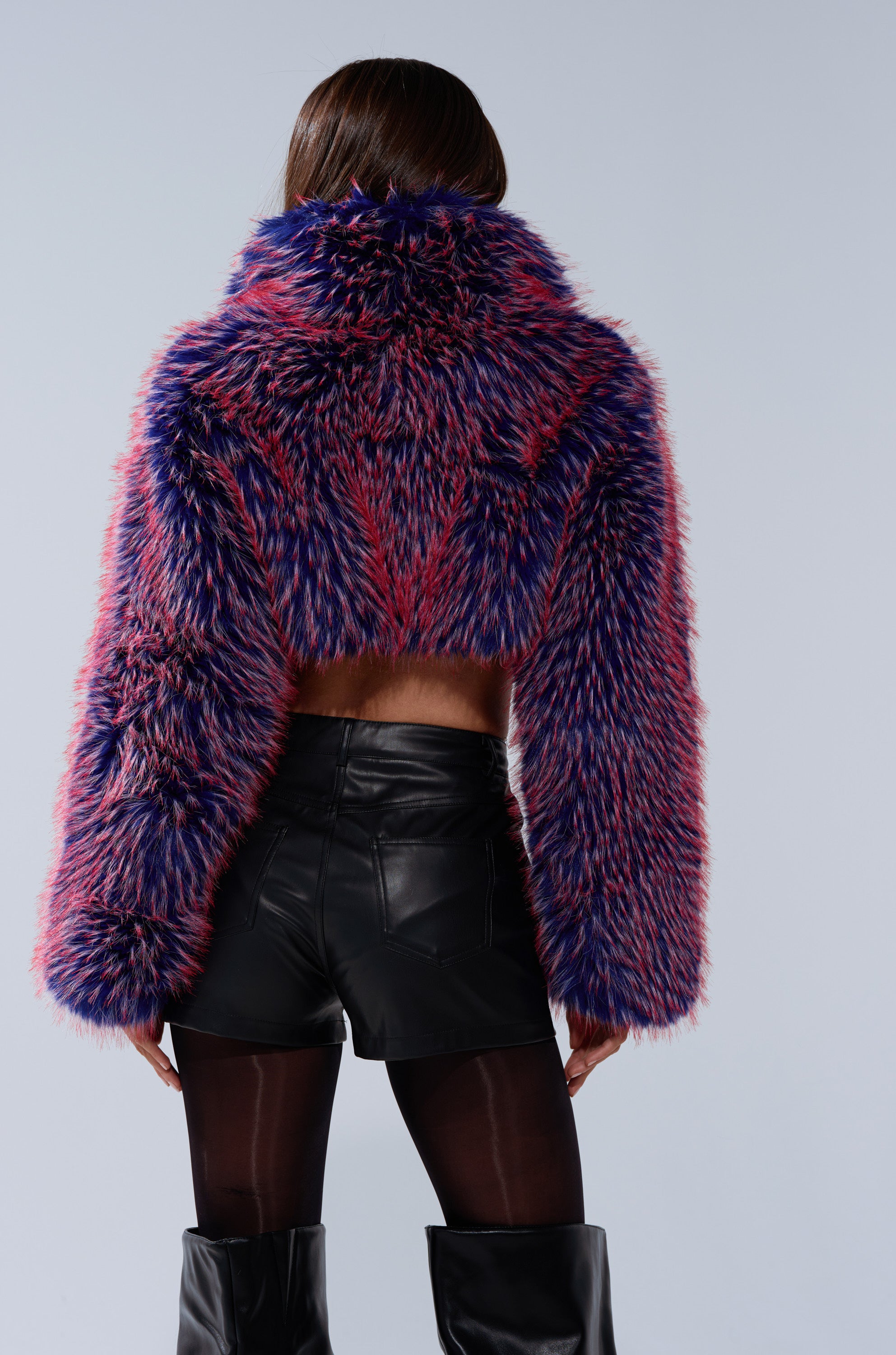 A person with long dark hair is seen from behind, wearing the NELLIE FAUX FUR BOLERO in purple, pink, and black, styled with black leather shorts, sheer tights, and tall black boots.