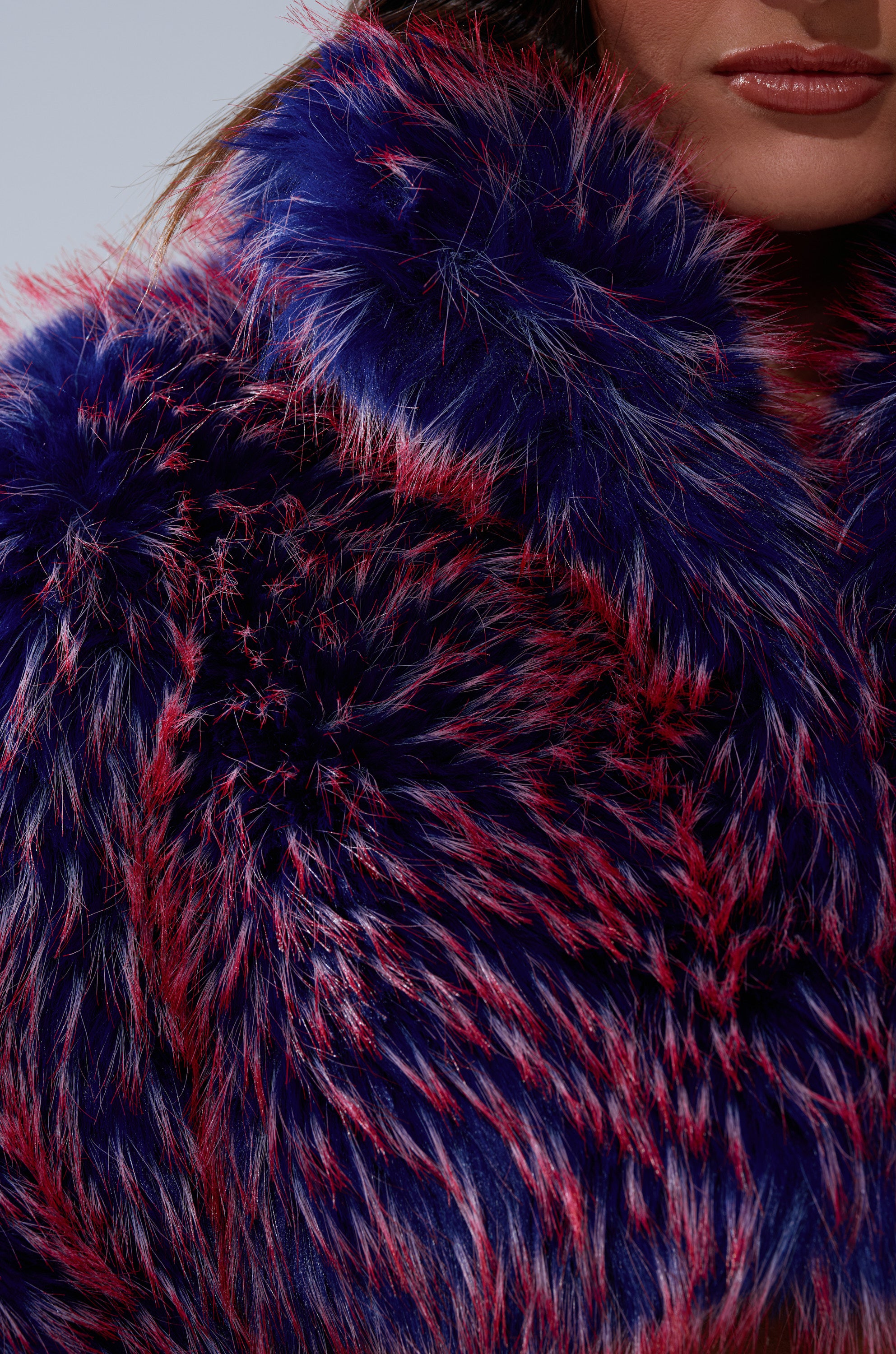 A close-up of a person wearing the NELLIE FAUX FUR BOLERO, featuring vibrant blue and red faux fur. Only the lower half of their face and neck are visible, emphasizing the jacket’s bold colors and fluffy texture.