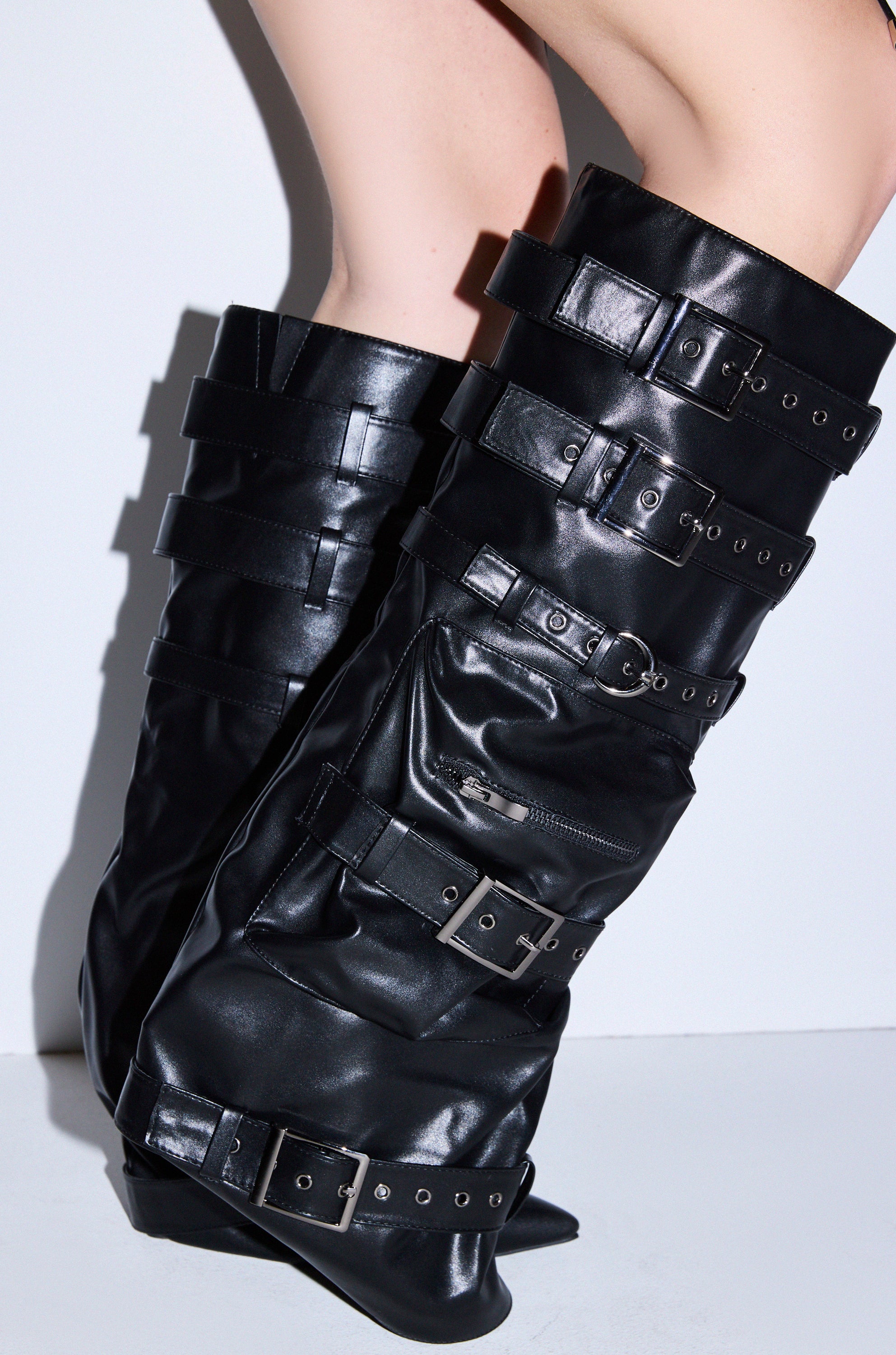 Close-up of a person's legs in AZALEA WANG NEMIANA BLACK BELT AND BUCKLES DETAIL BOOT—shiny black knee-high boots featuring multiple large straps, buckles, and zippers—standing on a white floor against a white background.