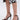 A person wears the AKIRA NEMTI BLACK STILETTO SANDAL with gold ankle straps and comfort cushions, standing on a light background. The hem of a black and white floral skirt is visible above the knees.