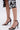 A person wears the AKIRA NEMTI BLACK STILETTO SANDAL with gold ankle straps and comfort cushions, standing on a light background. The hem of a black and white floral skirt is visible above the knees.