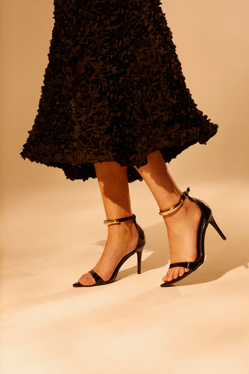 A person wearing a textured black skirt and the AKIRA NEMTI BLACK STILETTO SANDAL with ankle straps stands against a neutral background, with only their legs and part of the skirt visible.