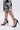 A person wearing a black and white floral skirt and chic AKIRA NEMTI BLACK STILETTO SANDAL with ankle straps and sleek patent leather upper stands on a light background.