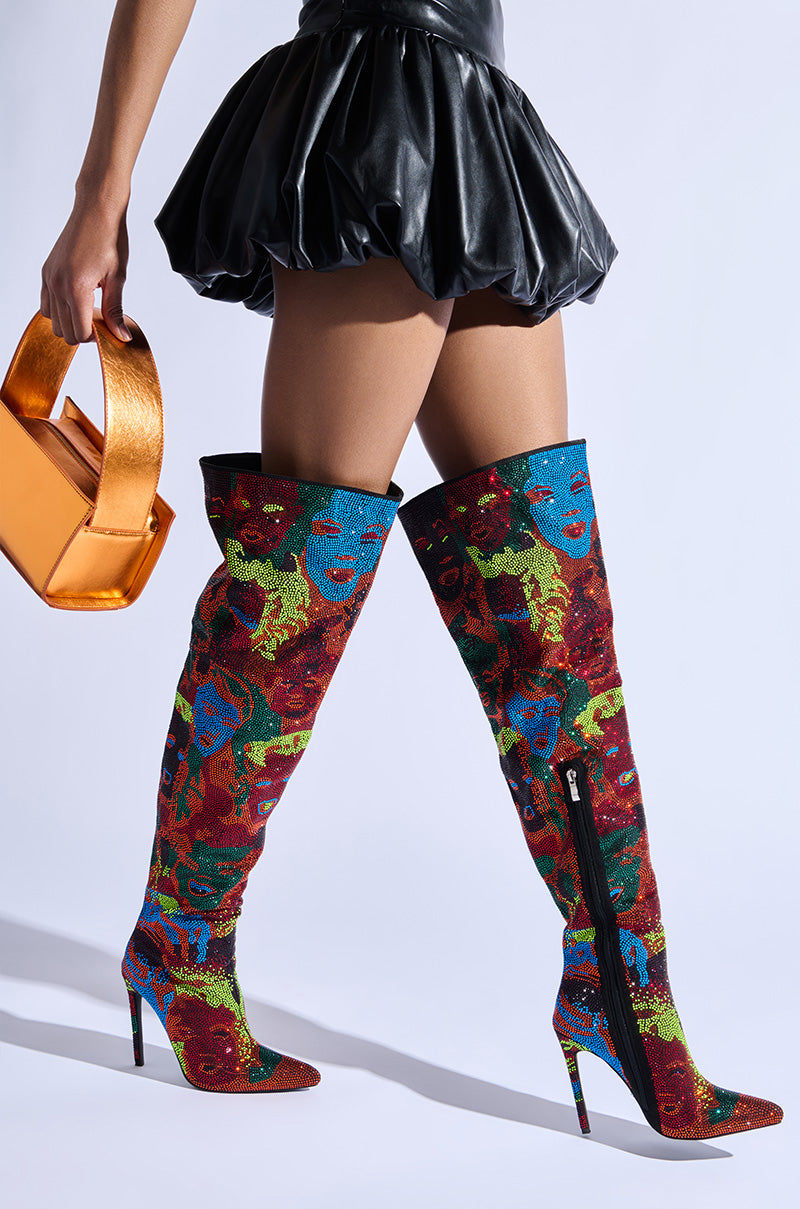 A person wearing a black leather mini skirt and the striking AZALEA WANG NERISSA MULTI STILETTO THIGH HIGH BOOTS, adorned with patterns of faces, holds a metallic orange handbag and struts with confidence.