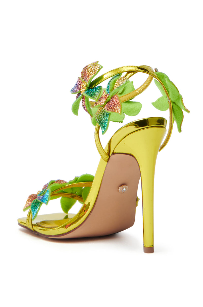 The AZALEA WANG NERIYA MULTI RHINESTONE FLOWER STILETTO SANDAL is a vibrant high-heel with a yellow-gold finish, featuring colorful butterfly embellishments, green leaf details, and ombre rainbow sequin shimmer on the straps. It showcases a slim stiletto heel and beige sole.