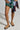 Against a light background, someone wears a mixed-media skirt with green and plaid patterns and denim. They showcase AZALEA WANG NESSAROSE tan faux suede sandals with flatform soles, along with a plaid long-sleeve shirt tied around the waist for an effortlessly chic look.