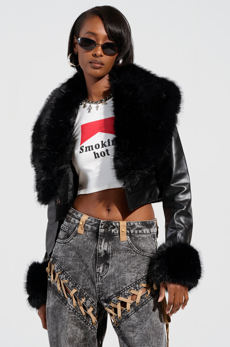 A person wears the eye-catching GISELE CROP MOTO BLAZER, a black leather jacket with fur trim, over a white crop top with text and gray jeans featuring crisscross lace details. The ensemble is finished with bold dark sunglasses and earrings against a plain backdrop.