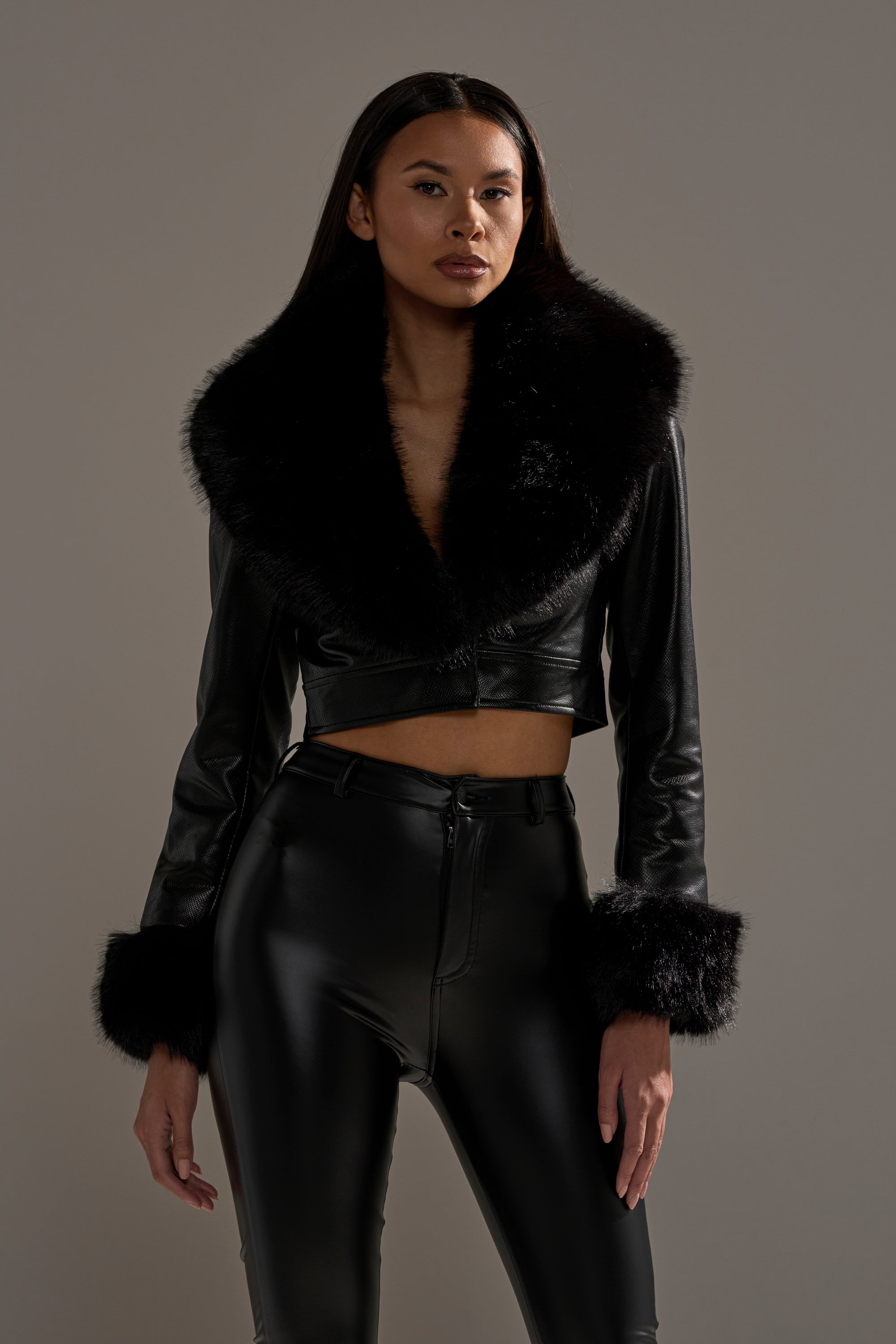 A woman stands against a neutral background wearing the GISELE CROP MOTO BLAZER styled with high-waisted black leather pants, her hands relaxed by her sides.