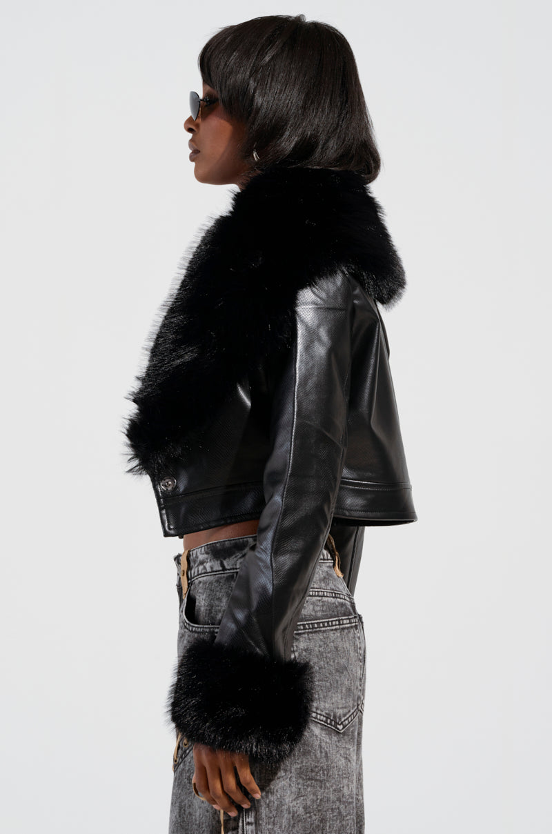 A woman wearing the GISELE CROP MOTO BLAZER, featuring faux fur trim on the collar and cuffs, is paired with high-waisted gray jeans. She has short hair and sunglasses while standing in profile against a plain white background.