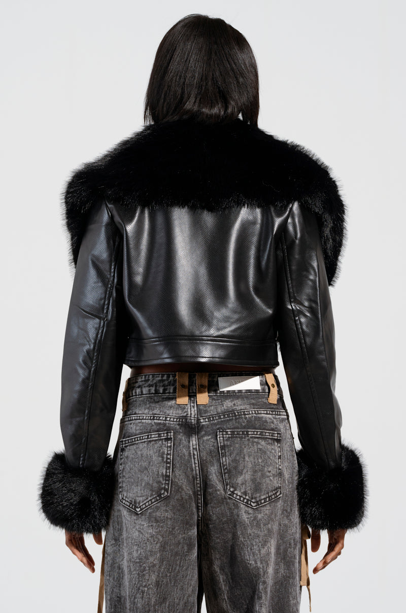 The rear view captures a person wearing the GISELE CROP MOTO BLAZER, a black croc-embossed faux leather jacket featuring a detachable faux fur collar and cuffs. They complement it with faded gray jeans and loosely hanging brown suspenders against a simple off-white canvas backdrop.