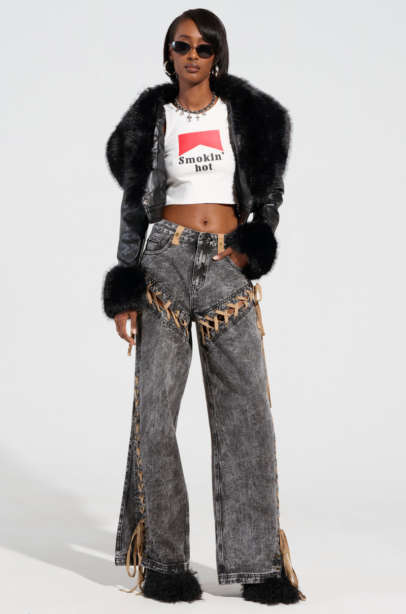 A woman wearing sunglasses, a stylish black GISELE CROP MOTO BLAZER with a detachable faux fur collar, and a white "smokin' hot" T-shirt, paired with grey wide-legged jeans featuring lace-up sides. She stands confidently against a plain white background.