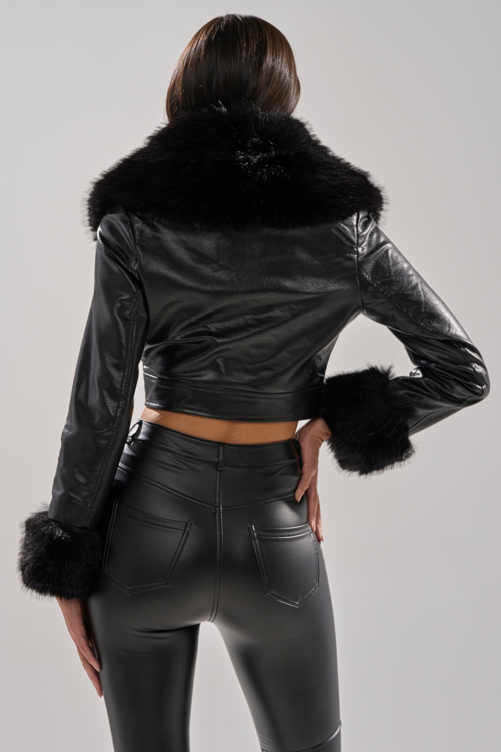 A person is shown from the back against a light background, wearing the GISELE CROP MOTO BLAZER, a black faux fur jacket with fur trim on the collar and cuffs, paired with high-waisted black faux leather pants.