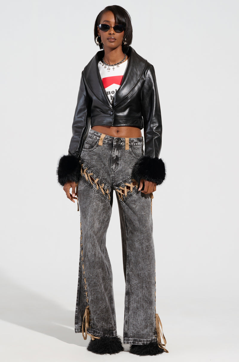 Sporting the GISELE CROP MOTO BLAZER, a black leather jacket with a detachable faux fur collar, this individual pairs it perfectly with a graphic t-shirt and high-waisted, wide-leg jeans featuring lace-up detailing. Their look is completed with sunglasses, straight black hair, and distressed jeans adorned with fur at the hems to match the jacket's stylish flair.
