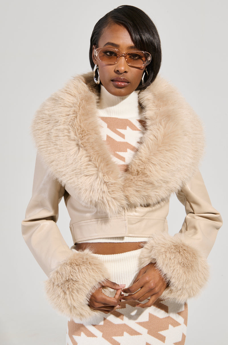 The woman dons sleek sunglasses and the Gisele Crop Faux Fur Moto Jacket in Ivory, featuring a chic, removable faux fur trim. Beneath, she wears a patterned beige and white sweater. Her stylish ensemble stands out against the plain background.