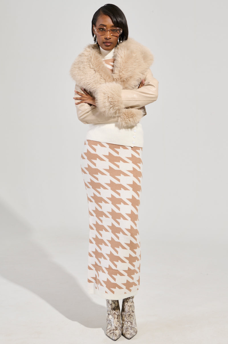 Dressed with assurance, an individual dons the GISELE CROP FAUX FUR MOTO JACKET IN IVORY, sunglasses perched stylishly. This jacket, featuring a removable faux fur trim, is layered over a dress with a houndstooth pattern and paired effortlessly with snakeskin-patterned boots against a simple, light-colored background.