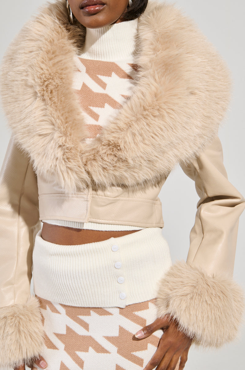 A person dressed in an ivory Gisele Crop Faux Fur Moto Jacket layers it over a houndstooth-patterned top and coordinating skirt, showcasing white ribbed trim and button accents, along with detachable faux fur trim on the cuffs. The person's face is not visible.