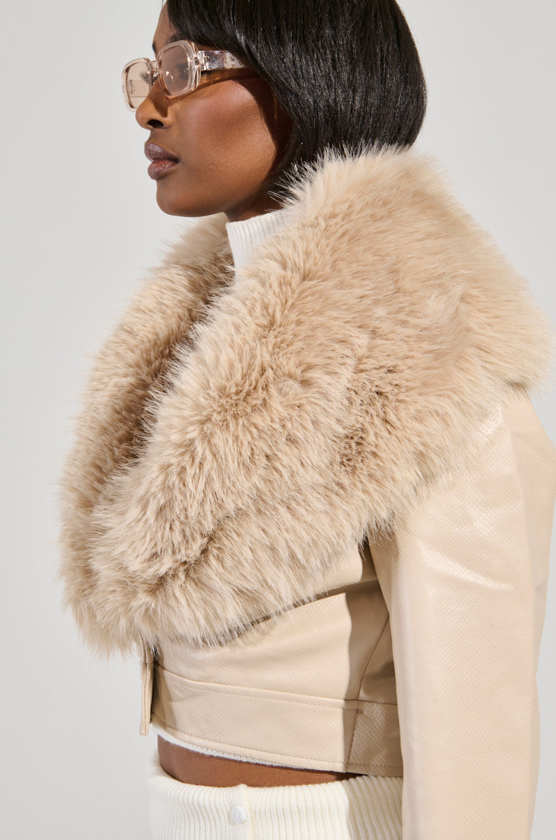 A person wearing clear glasses and a light beige jacket styled as the Gisele Crop Faux Fur Moto Jacket in Ivory. The jacket includes a removable faux fur trim over a white turtleneck, with their side profile captured against a neutral background.