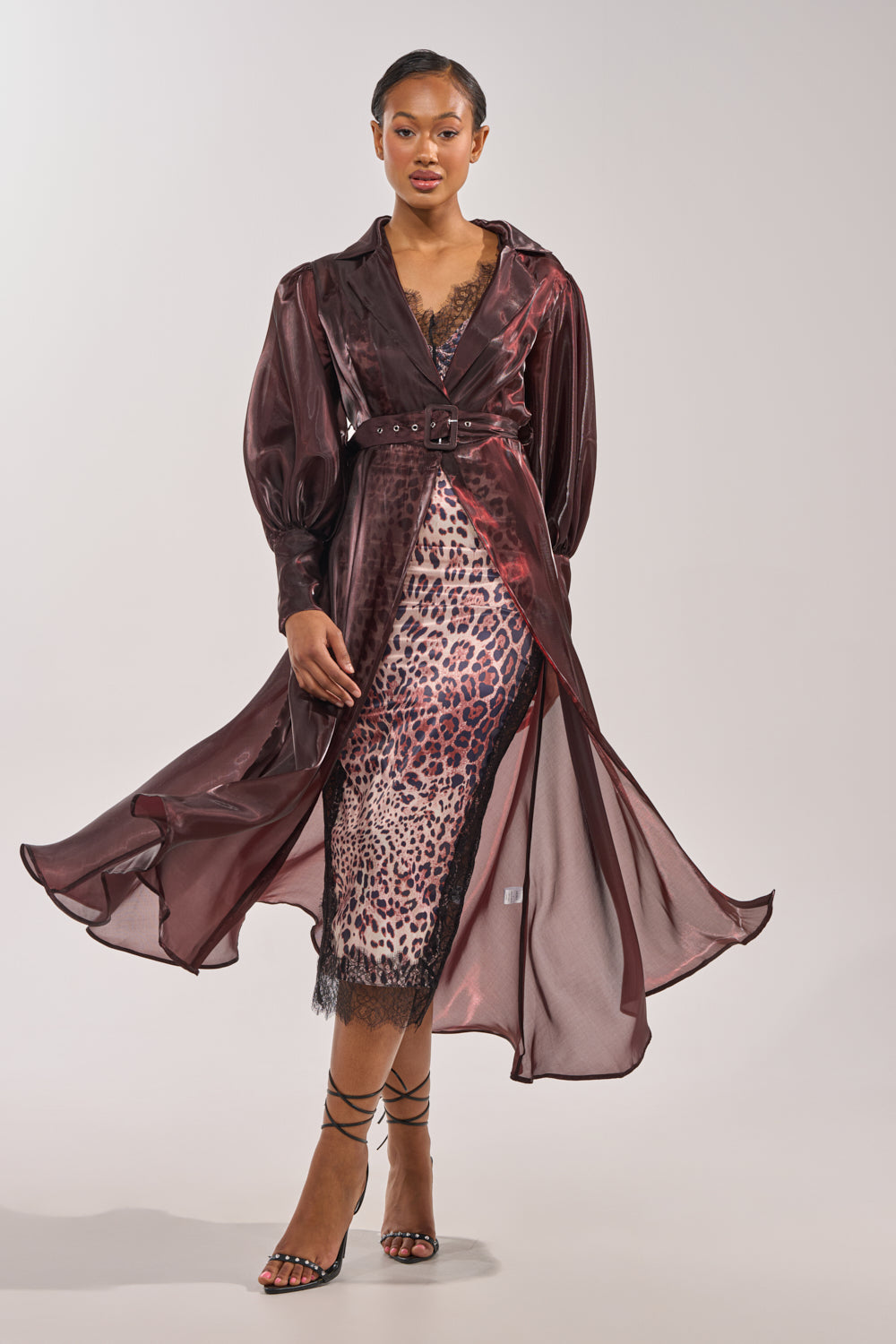 A woman poses confidently in the PRETTY LIL THING ORGANZA TRENCH, layered over a lace-trimmed leopard print dress and strappy black heels, exuding elegance against a simple light backdrop. 0