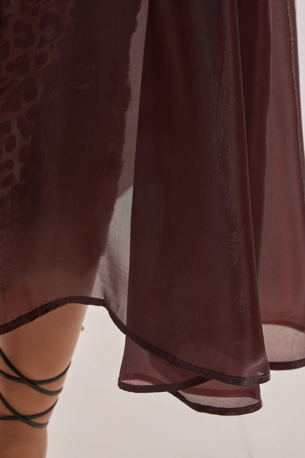 A close-up of the PRETTY LIL THING ORGANZA TRENCH—a sheer brown trench coat—styled over a sheer burgundy skirt with a wavy hem. Leopard-print fabric subtly peeks through, and black strappy sandals complete the look. 9