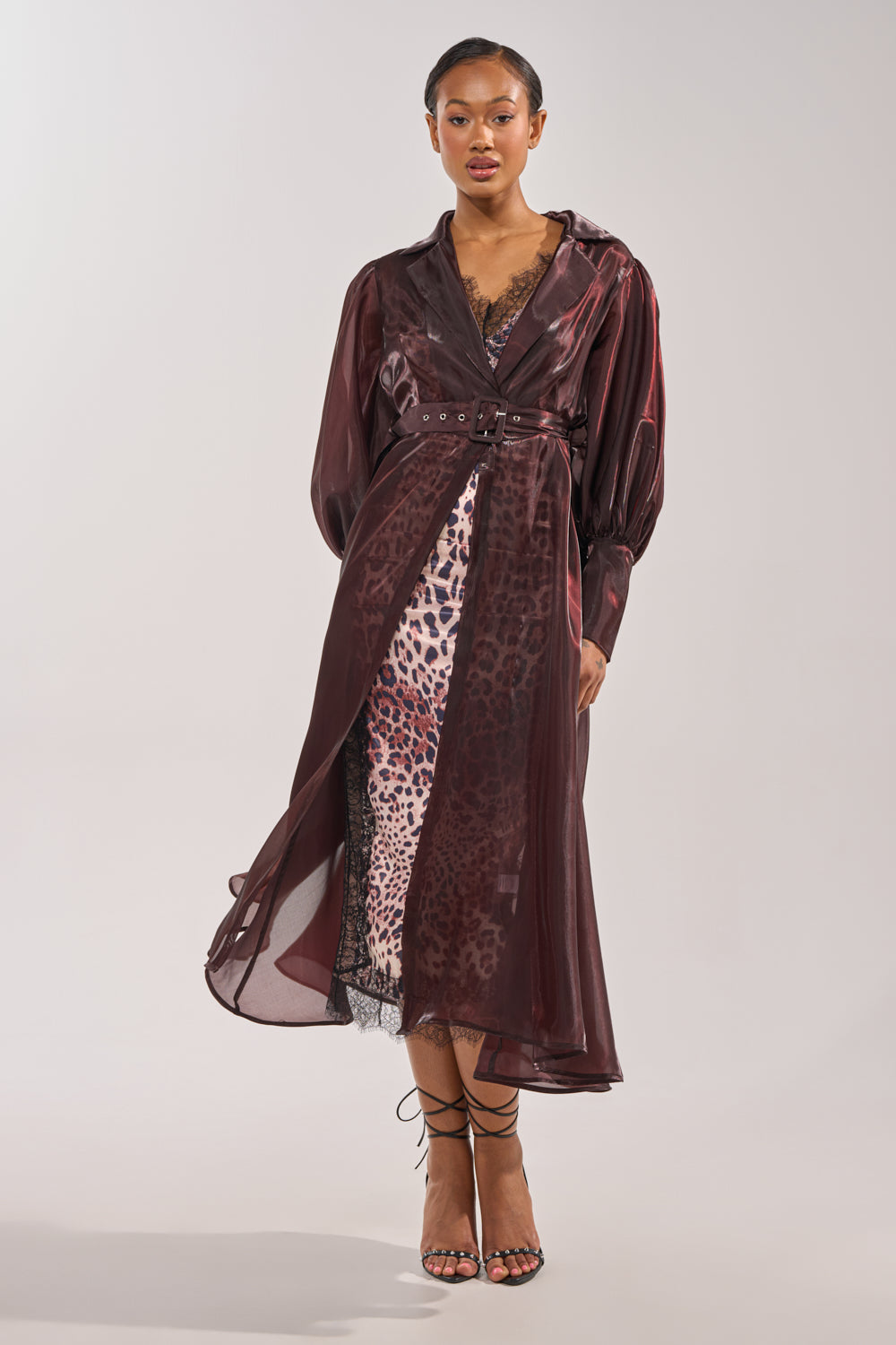 A woman wears the PRETTY LIL THING ORGANZA TRENCH, a sheer brown coat, over a leopard print dress with lace trim and strappy sandals, standing forward against a plain light background to showcase chic outerwear. 1