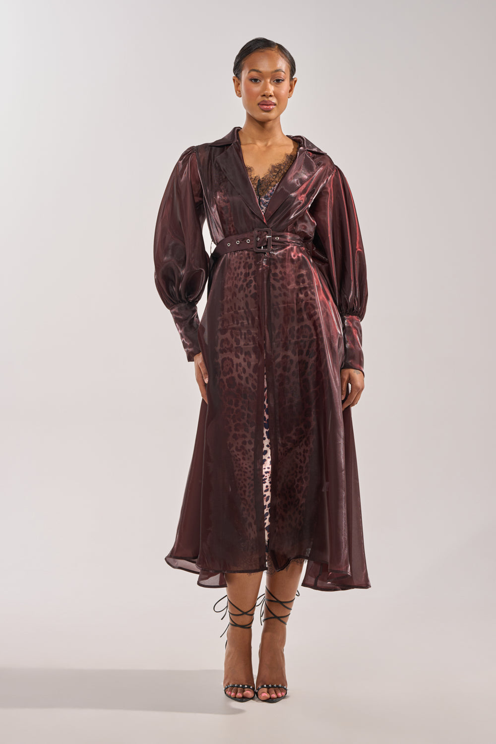 A woman faces forward in the PRETTY LIL THING ORGANZA TRENCH, a sheer brown belted coat layered over a leopard-print slip. She wears strappy heeled sandals, her hair slicked back, set against a plain background. 2