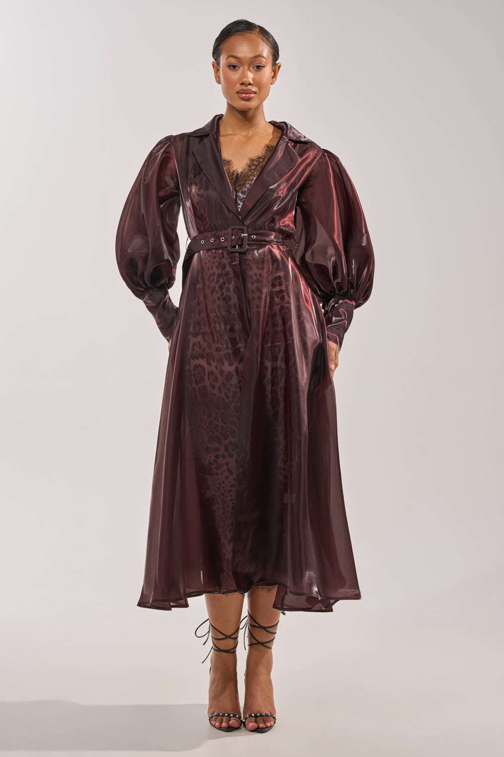 A woman wears the PRETTY LIL THING ORGANZA TRENCH, a shiny, deep burgundy belted coat dress with puffed sleeves and a subtle leopard print, styled with strappy black heels and her hair neatly pulled back. 3